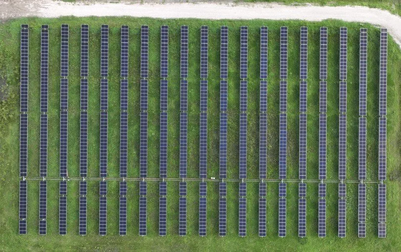 Aerial view of a solar panel farm with multiple rows of blue solar panels on green grass, bordered by a gravel path.