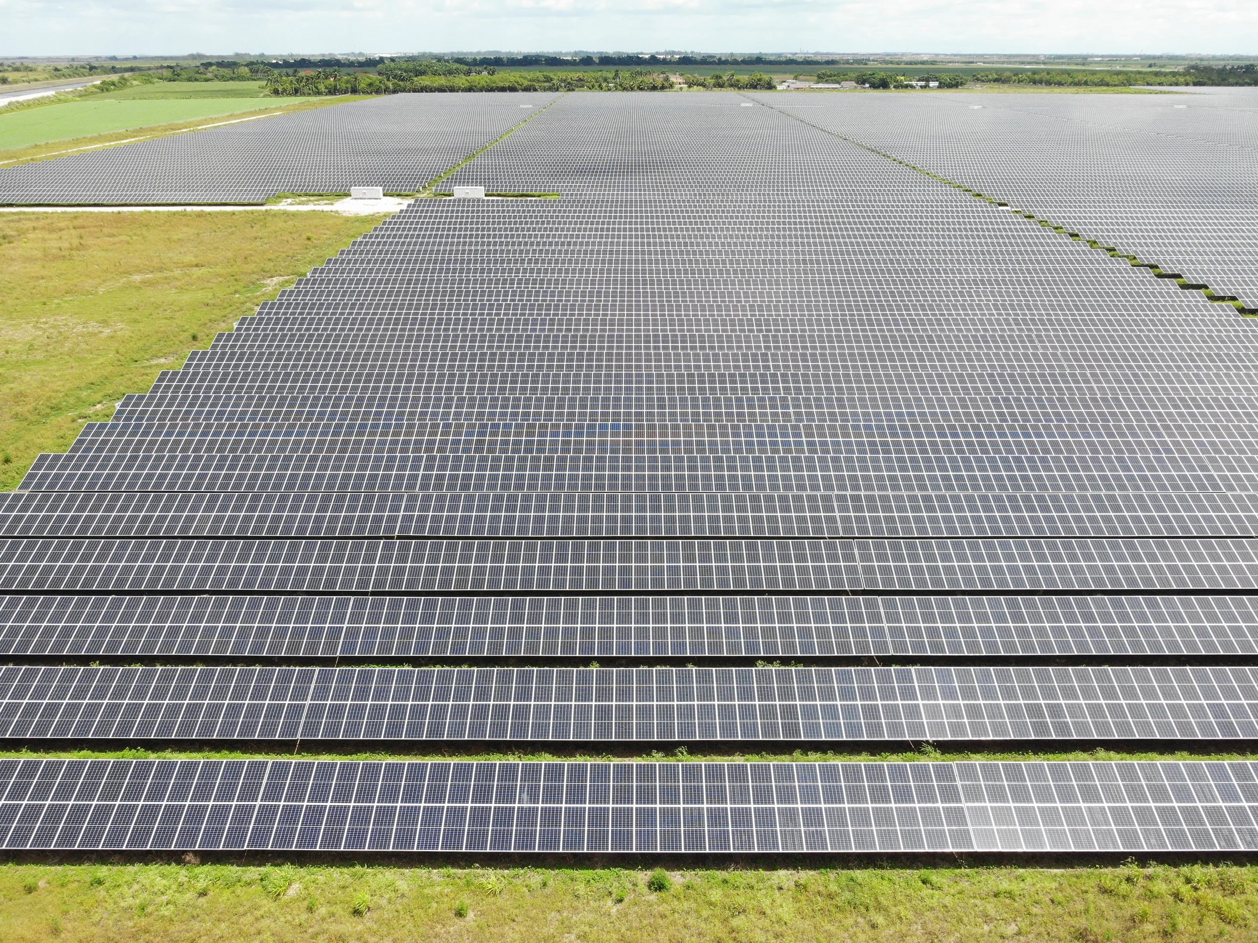 An expansive solar farm with rows of solar panels on a sunny day, surrounded by green fields and distant trees.