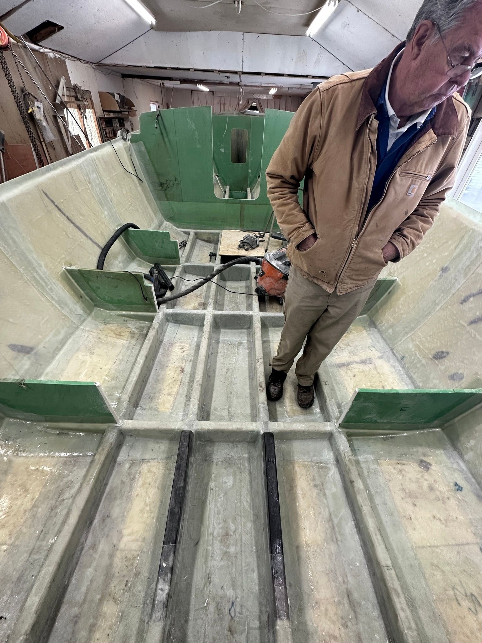    Bruce Dyson stands in the boat with a fully fiberglassed interior hull, the hull structural framework complete, and forward bulkheads and deck supports in place, March 2023   