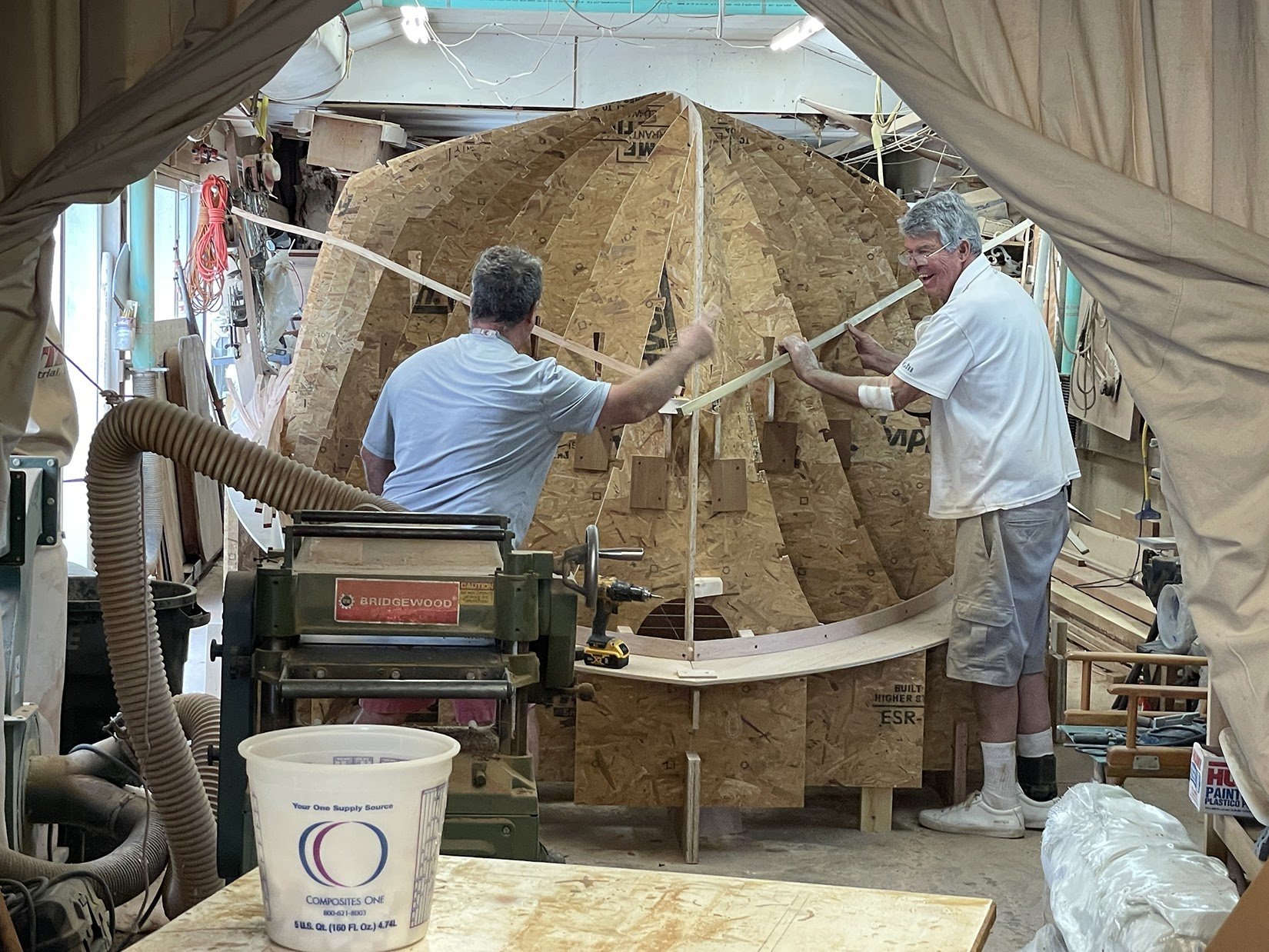    Andrew Flaherty (L) and Bruce Dyson (R) add strips of wood to the plywood “strongbacks” to better define the shape of the hull before foam Corecell is applied, August 2022   