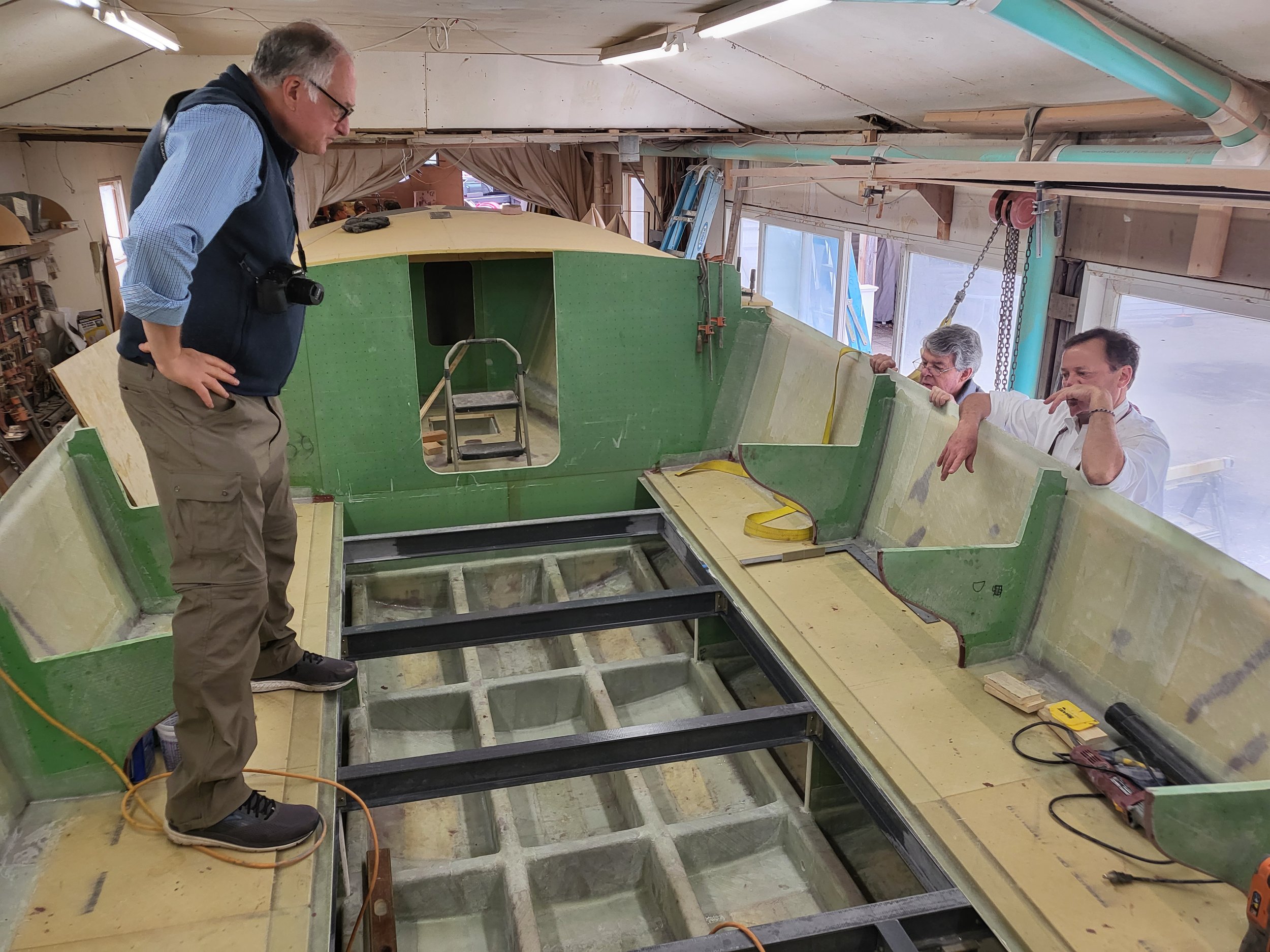    Neil Burns stands on the port (left) side of the future cabin deck straddling a passenger bench support while discussing things with Bruce Dyson and Doug Zurn.&nbsp; Note the Corecell foredeck in place awaiting fiberglass, and the fiberglass I-bea