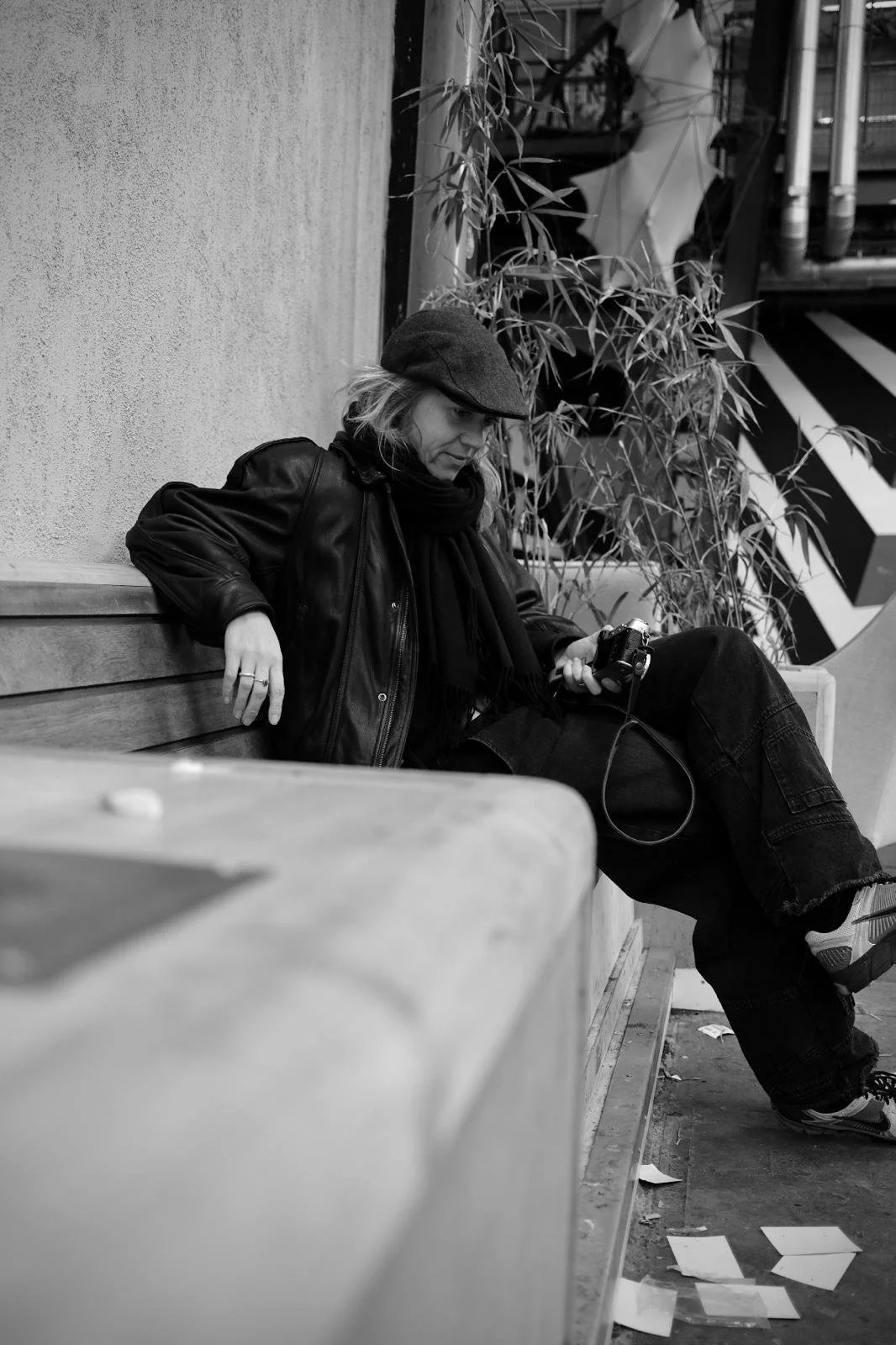 A woman sitting on a bench outdoors, looking at a camera in her hand, dressed in a leather jacket, scarf, and hat.