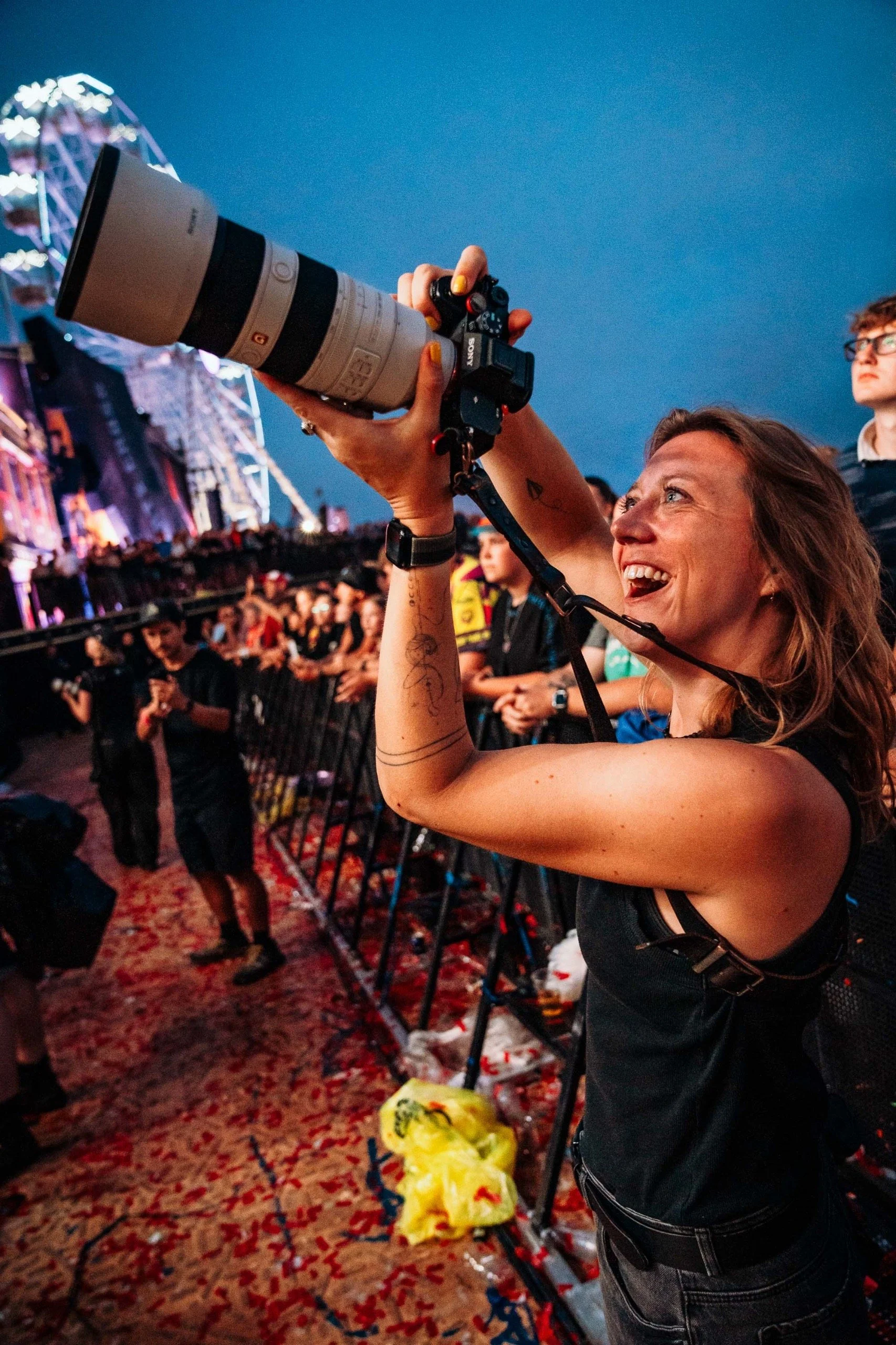 A woman at a concert taking a photograph with a professional camera with a large telephoto lens. There are other concert attendees and a stage with colorful lighting in the background.
