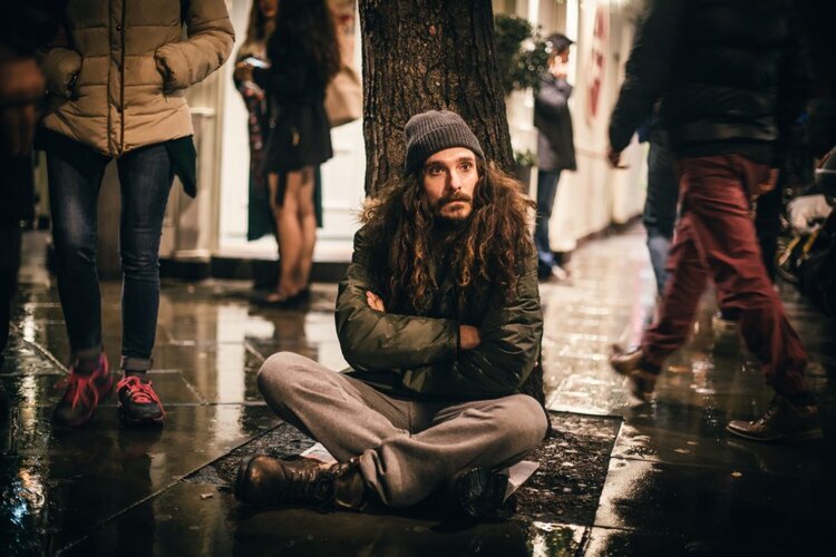  bearded man sits with folded arms under a tree as strangers walk past 