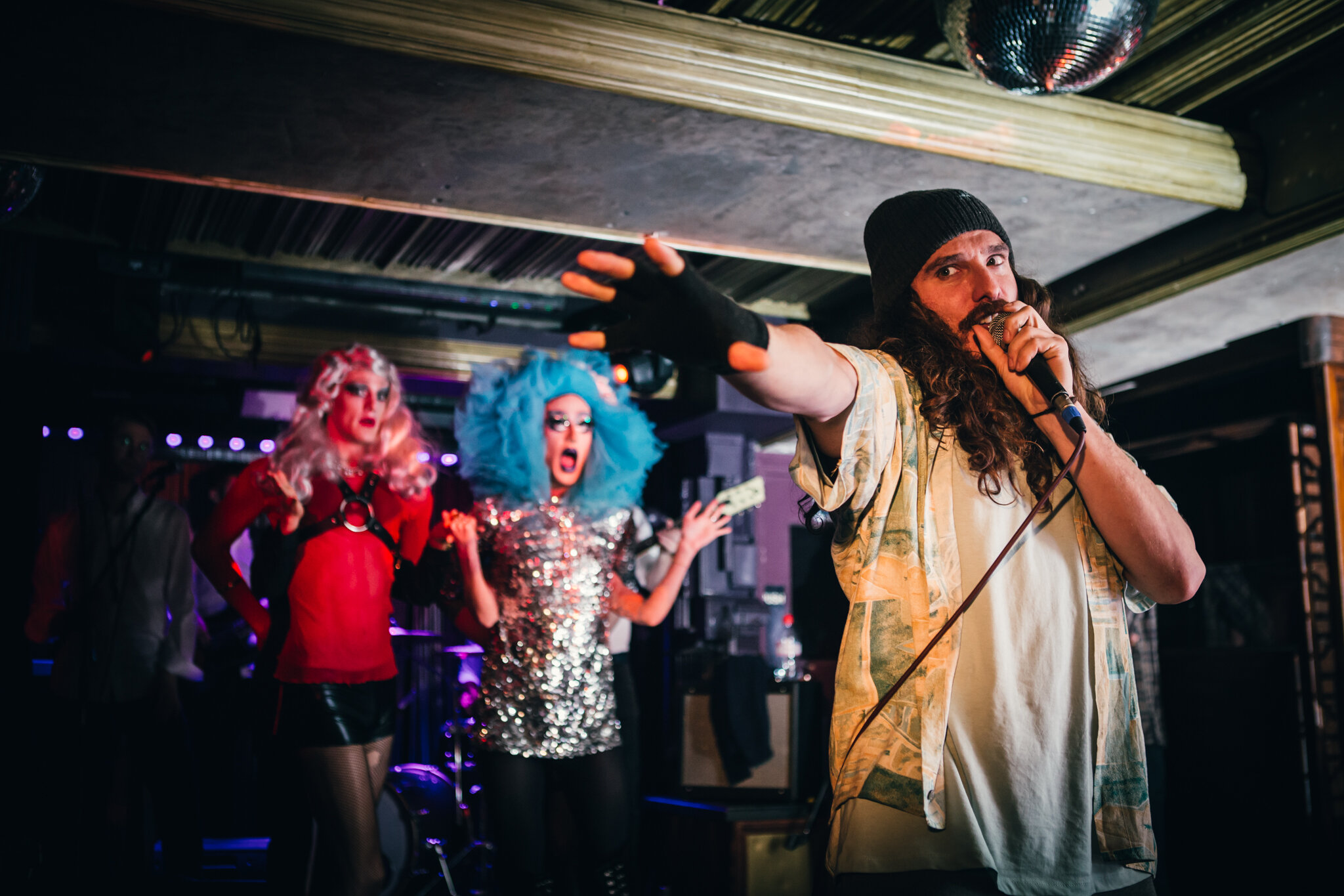  bearded man singing into microphone in front of two drag queens with a band in a club 