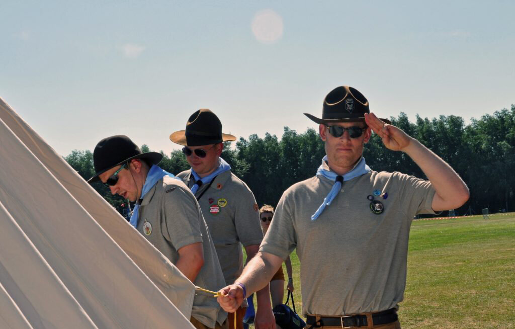  scouts in uniform saluting constructing tent outdoors on sunny day 
