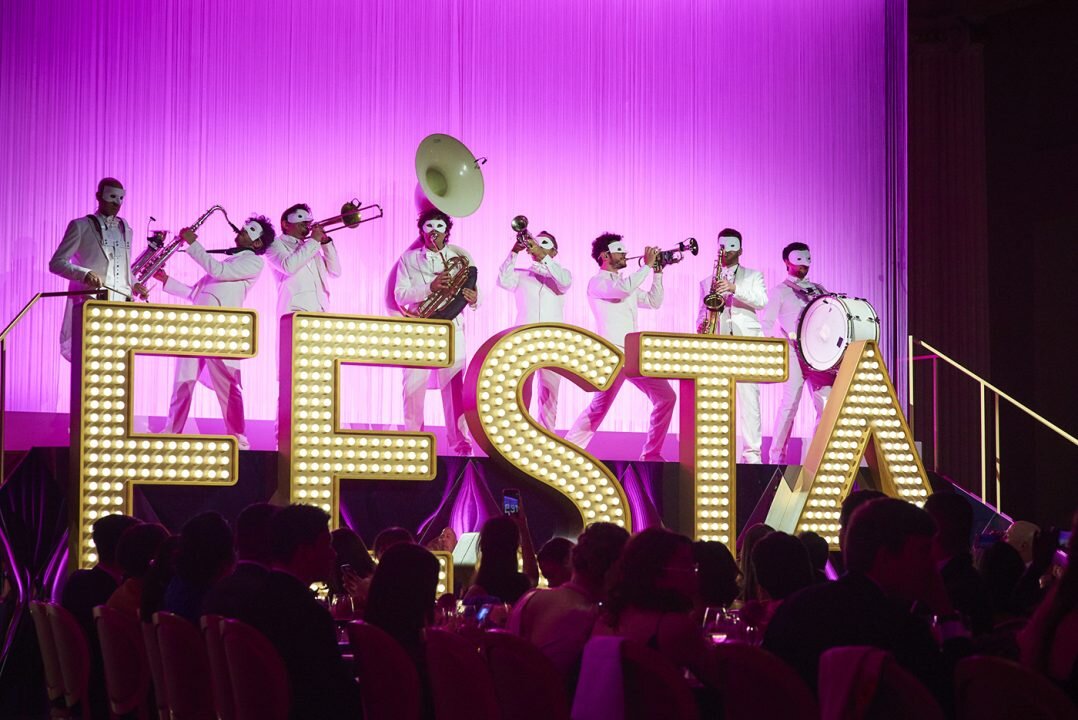  masked band dressed in white playing instruments on stage against pink backdrop and festa sign 