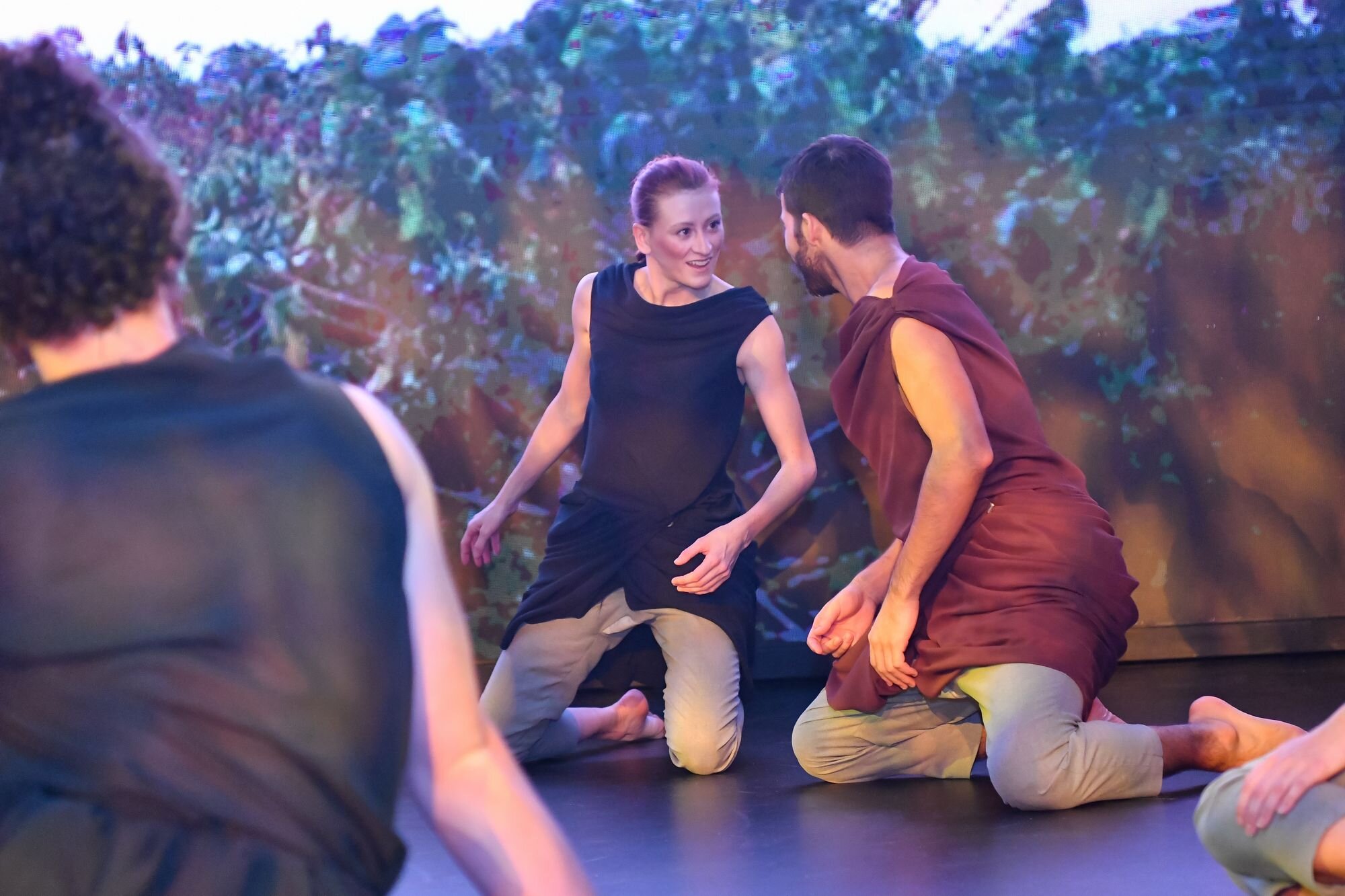  A male dancer and a female dancer wearing tunics kneel on the ground, engaging playfully with each other as others look on. 