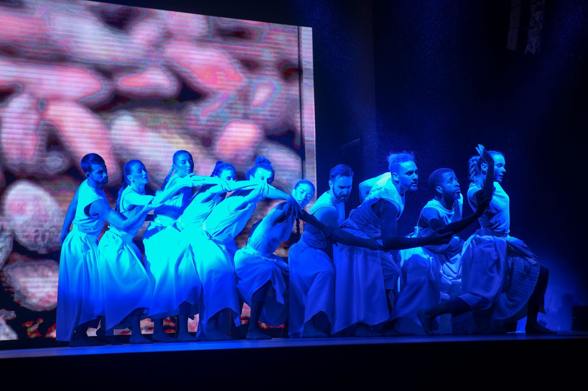 10 dancers create a wave of motion with their bodies, captured in a moody blue lighting state. 