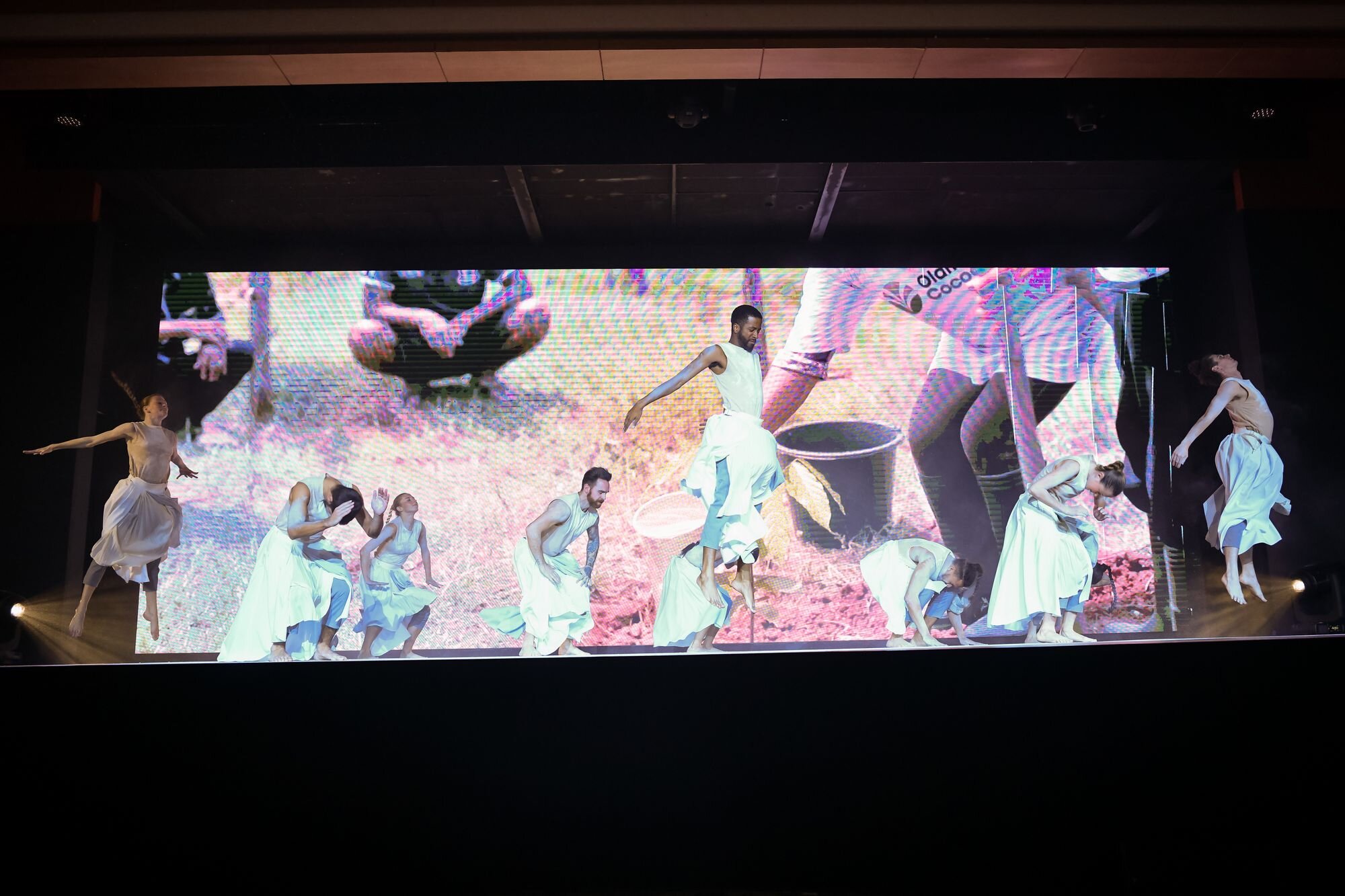  10 dancers dressed in flowing white garments are placed across a wide stage in front of a screen showing images of earth. They are in various states of jumping high into the air. 