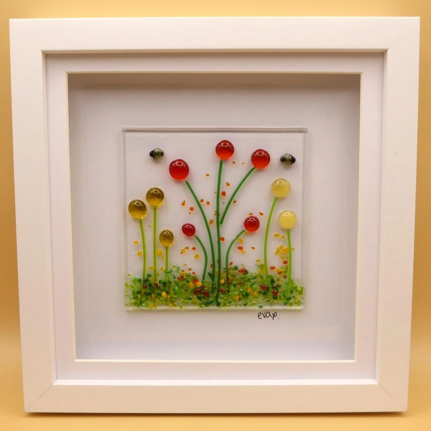 Fused glass artwork in white frame featuring bees flying above a wildflower meadow with orange and yellow flowers.  
