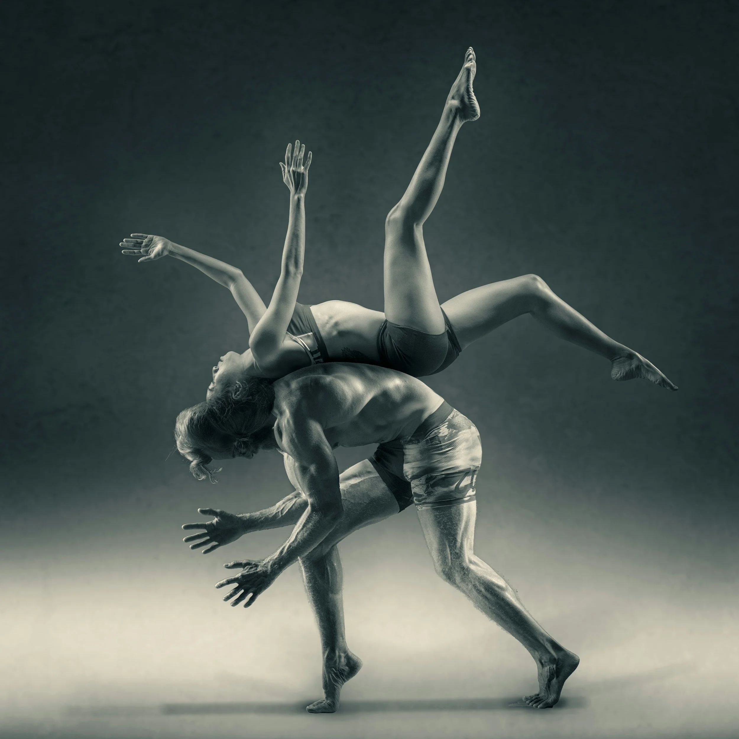 Two performers engaged in an acrobatic dance pose, with one balancing on the other's back, both with muscular builds and focused expressions.