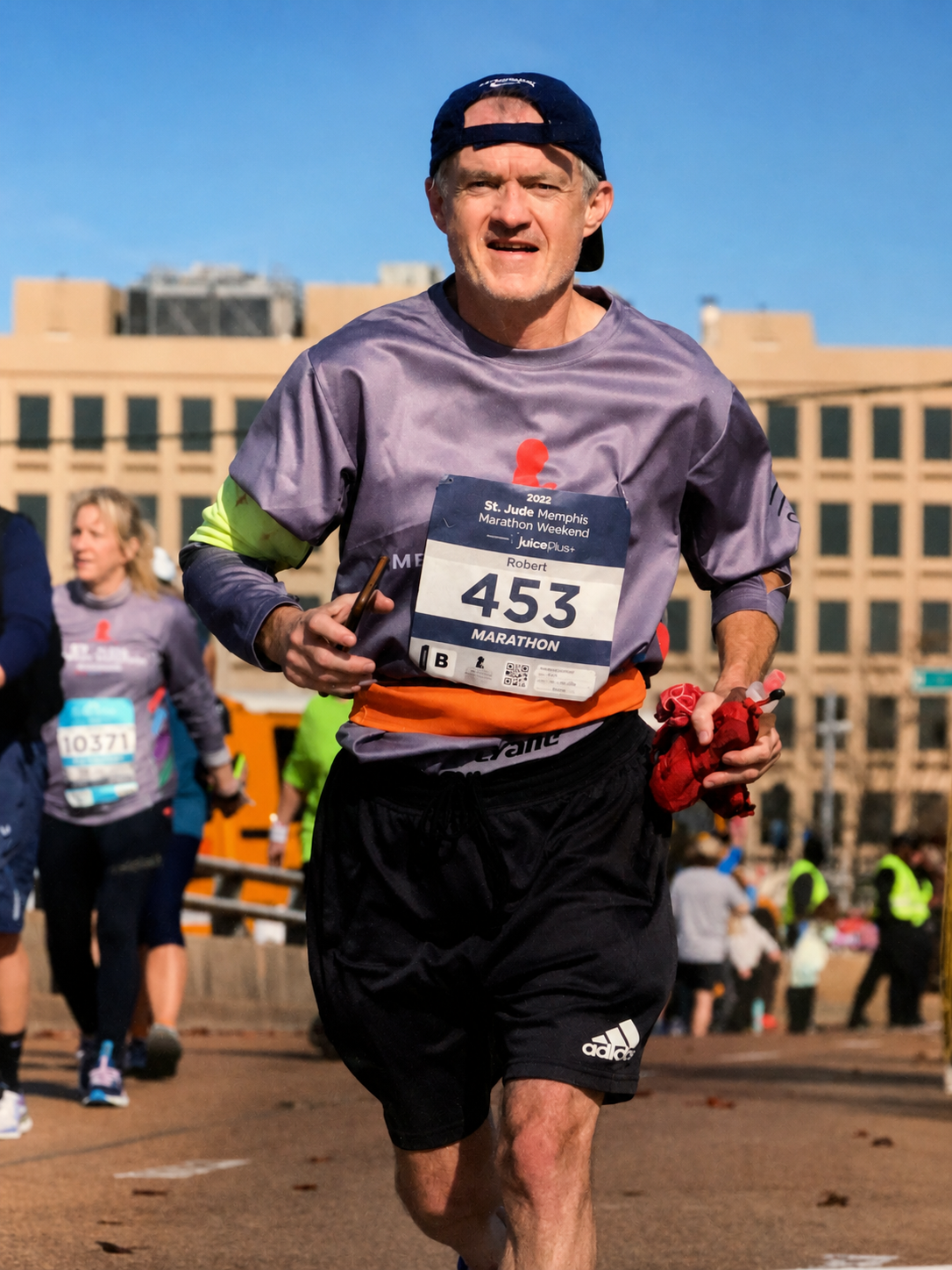 A man running in a marathon, wearing a purple shirt, black shorts, and a blue cap backward, with a yellow sleeve visible on his left arm. He is holding a red cloth in his left hand and appears to be near the finish line, with other runners and a cityscape in the background.