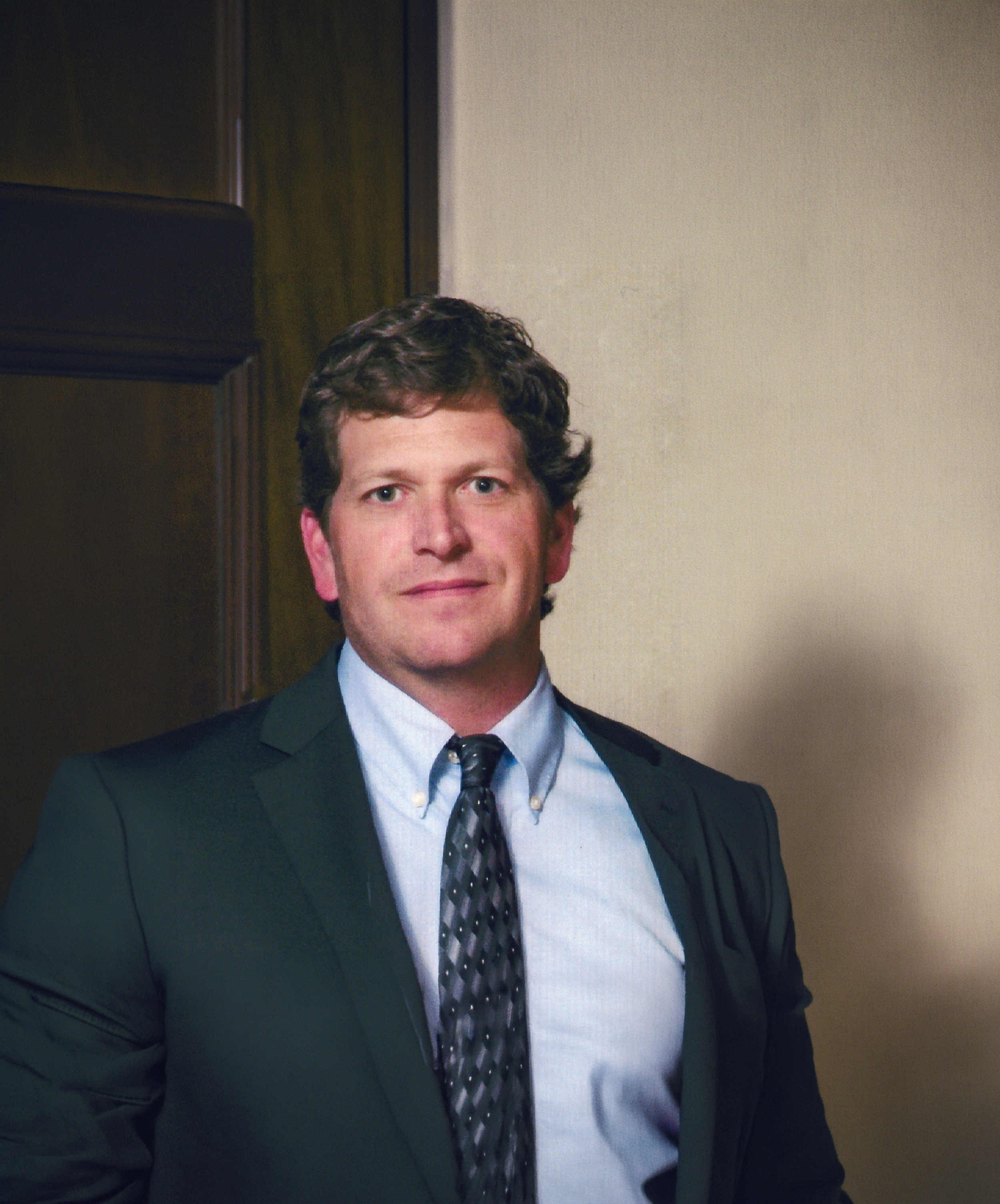 A man in a suit, tie, and collared shirt, standing indoors against a plain wall and wooden door, with only the top half of his face visible.