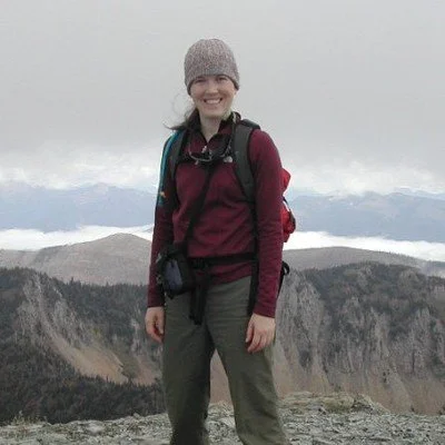 A woman smiling while hiking in a mountainous landscape with cloudy skies.