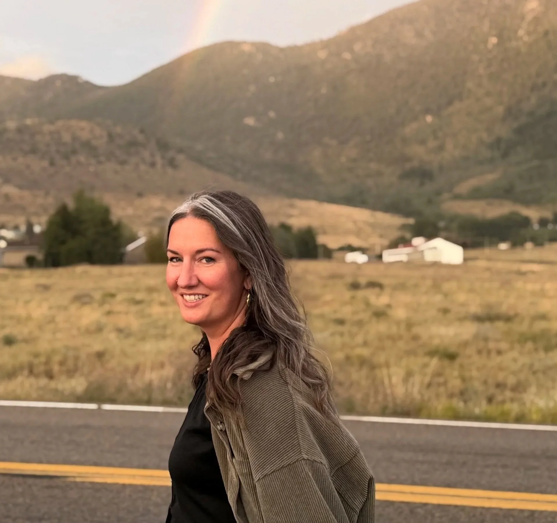 A woman with long gray hair and earrings smiling outdoors with a mountain and field in the background.