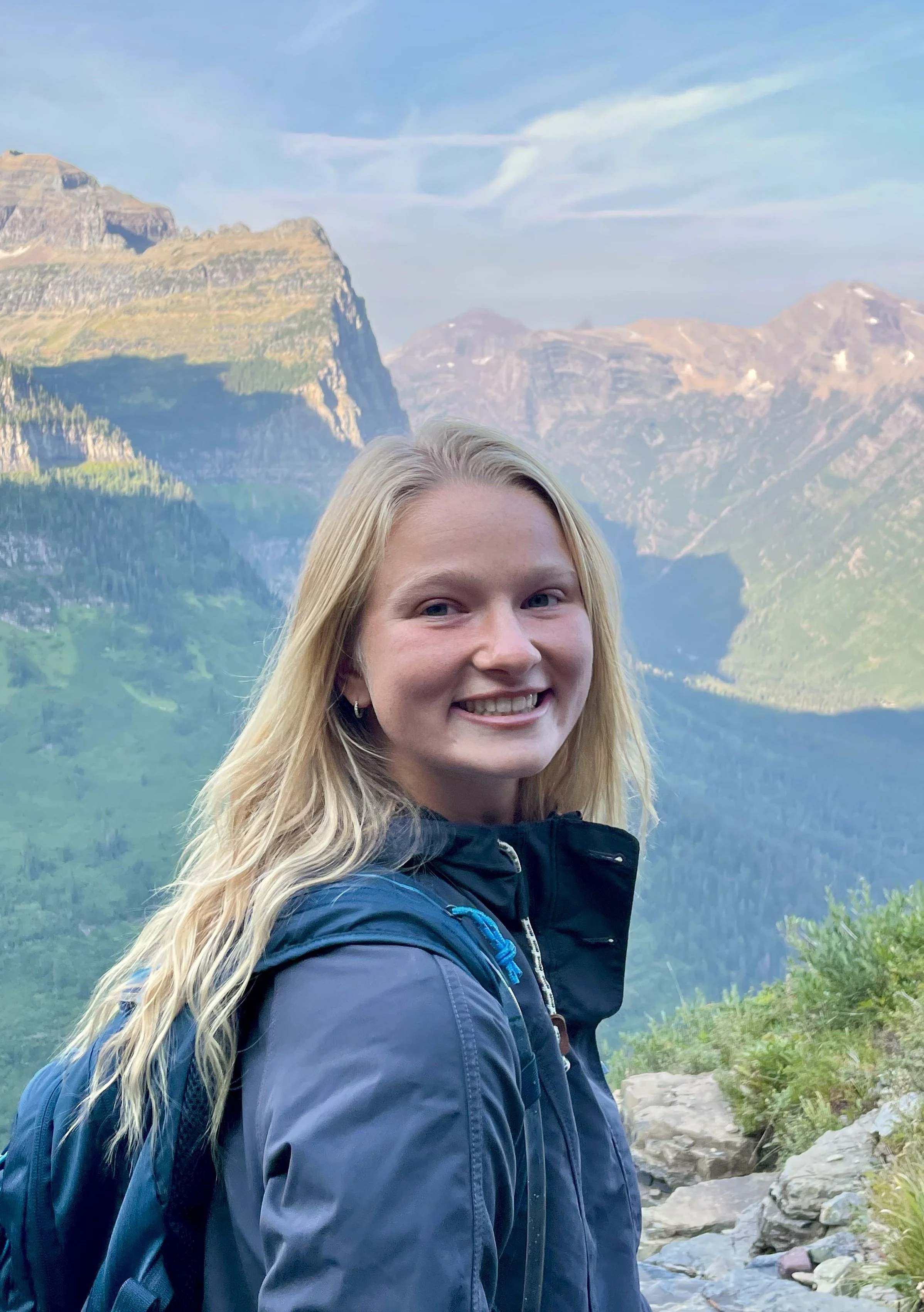 A smiling woman with long blonde hair wearing a blue jacket and carrying a blue backpack, standing outdoors in a mountainous landscape with steep cliffs, green trees, and a rocky stream.