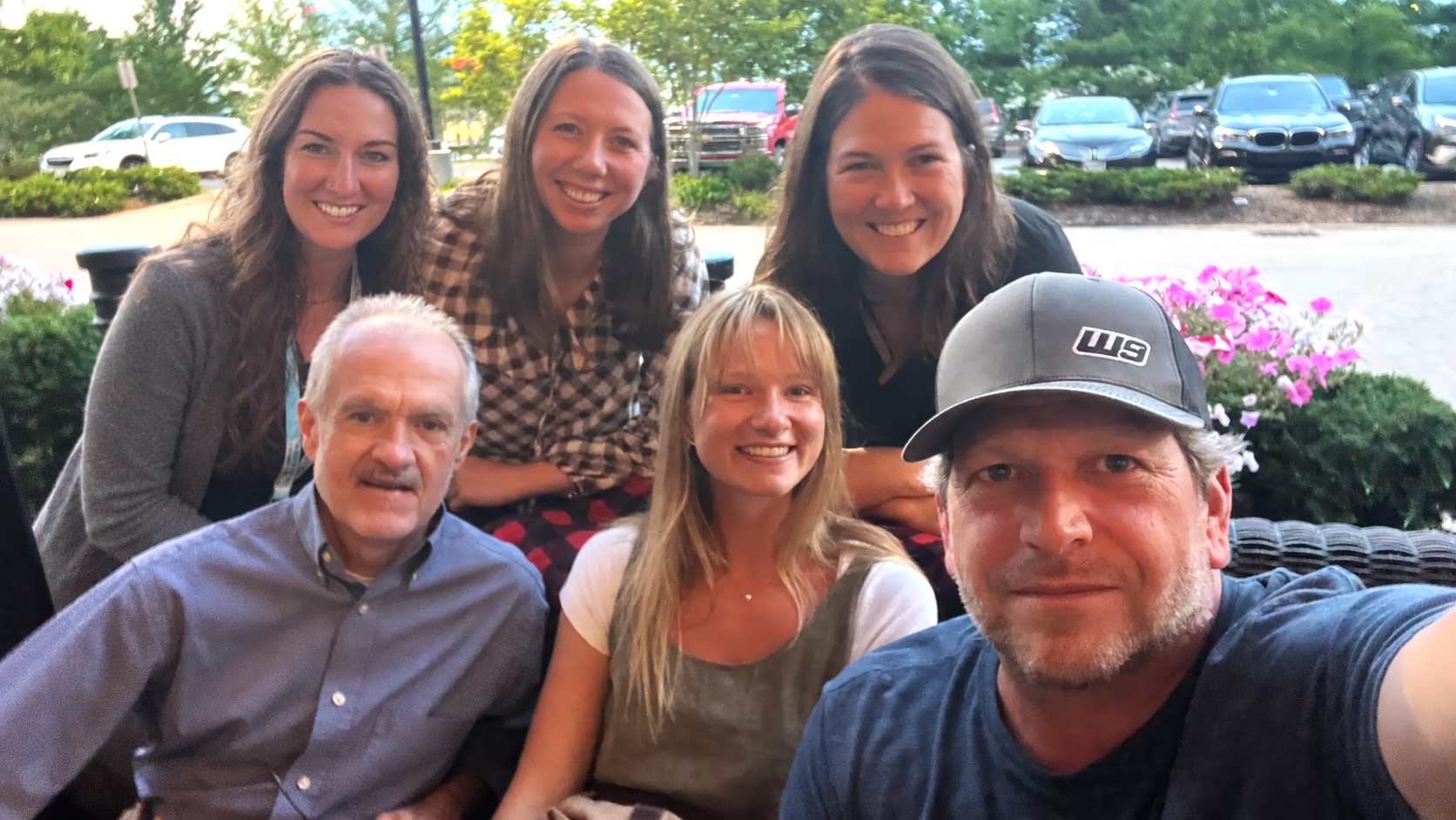 Group of six people smiling at a restaurant or cafe outdoors with car parking and blooming flowers in the background.