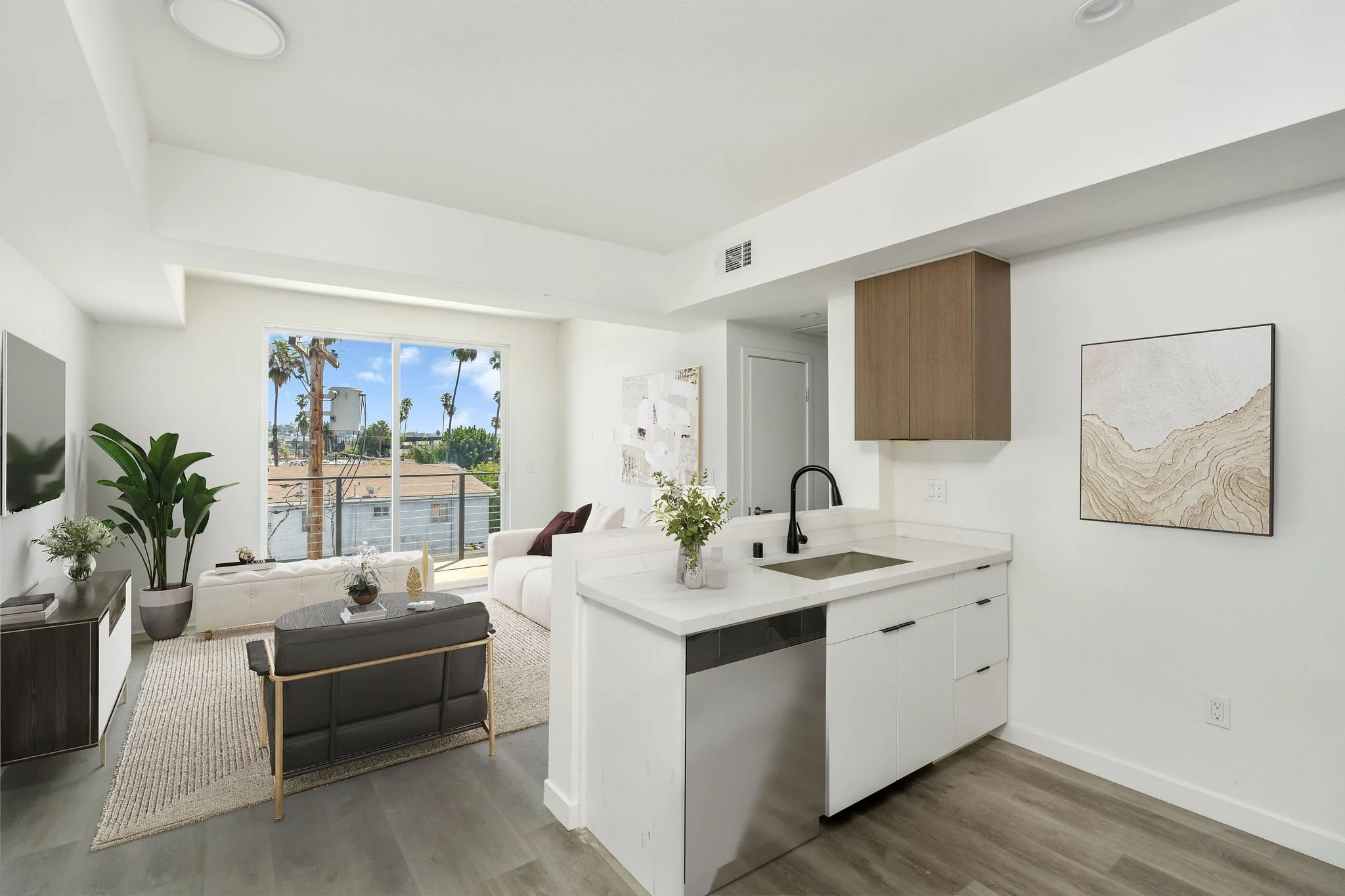 Open-concept living room and kitchen with large window view of palm trees and blue sky, modern white couch and a dark armchair, minimalist decor, and a sliding glass door leading to a balcony.