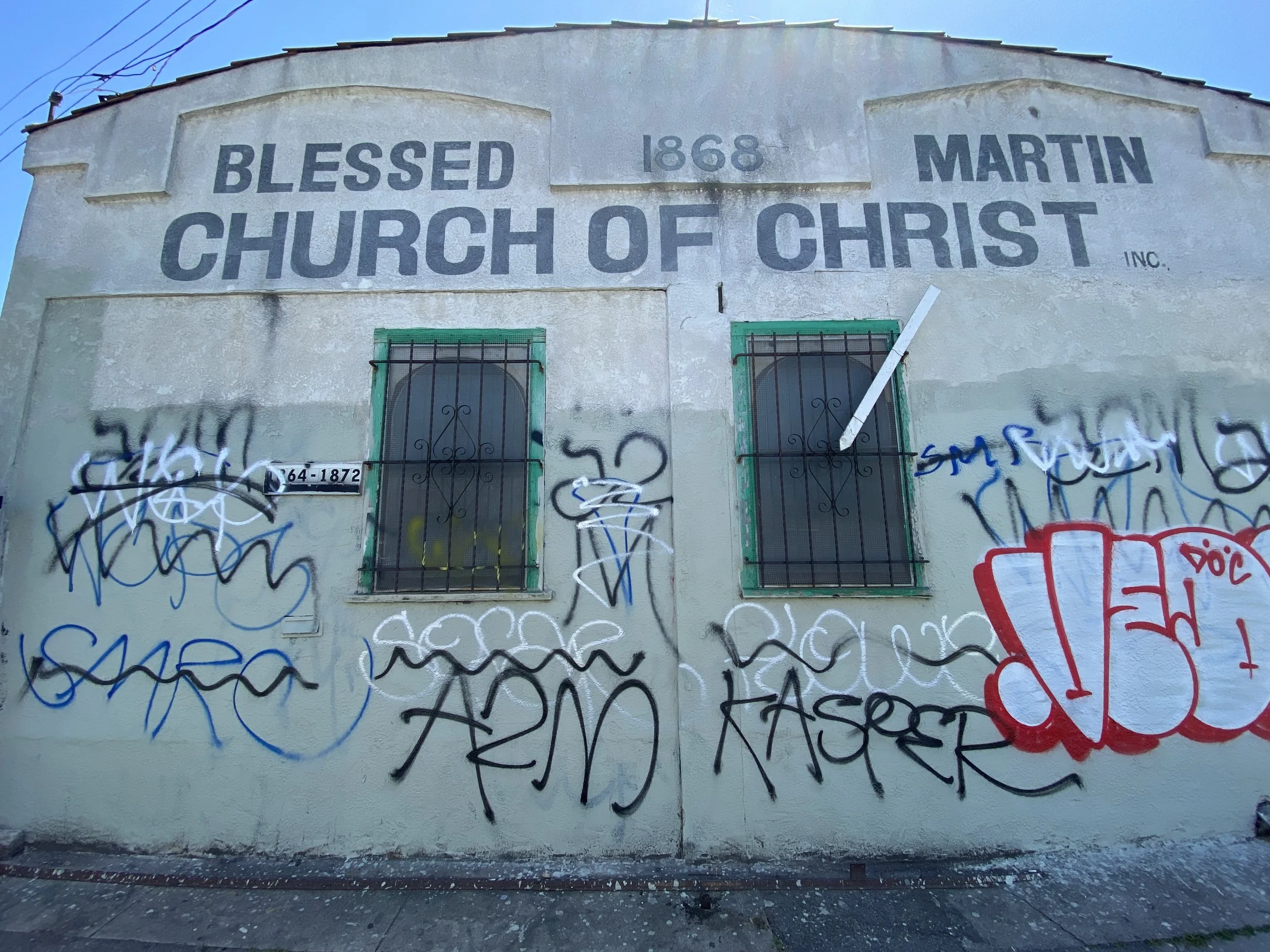 The exterior wall of the Blessed Martin Church of Christ, established in 1868, featuring two barred windows with graffiti and a faded church name on a light-colored wall.