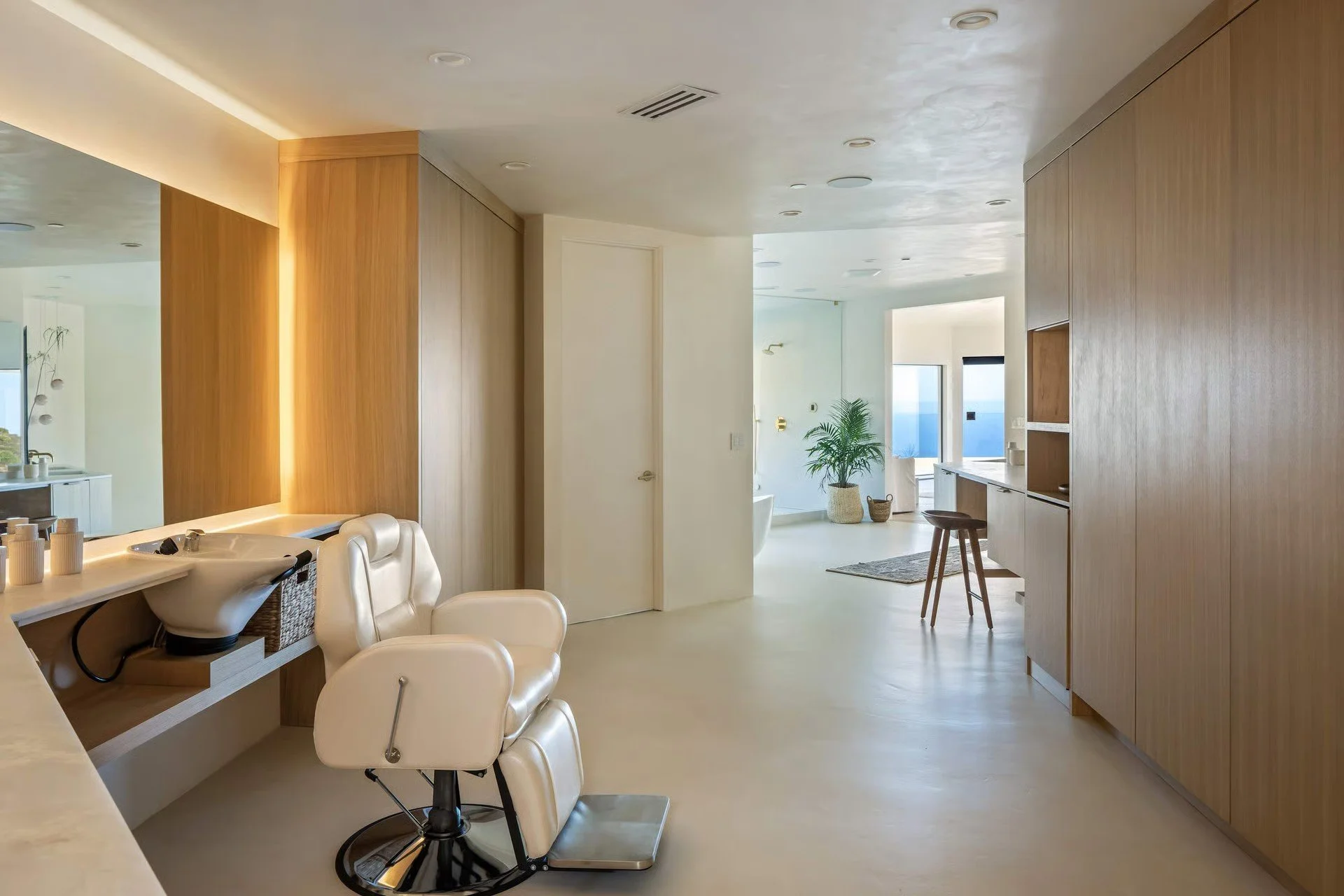Modern, light-colored living space with wooden and white finishes, a large window showing the ocean, a green potted plant, leather salon chairs, and a small dining area.
