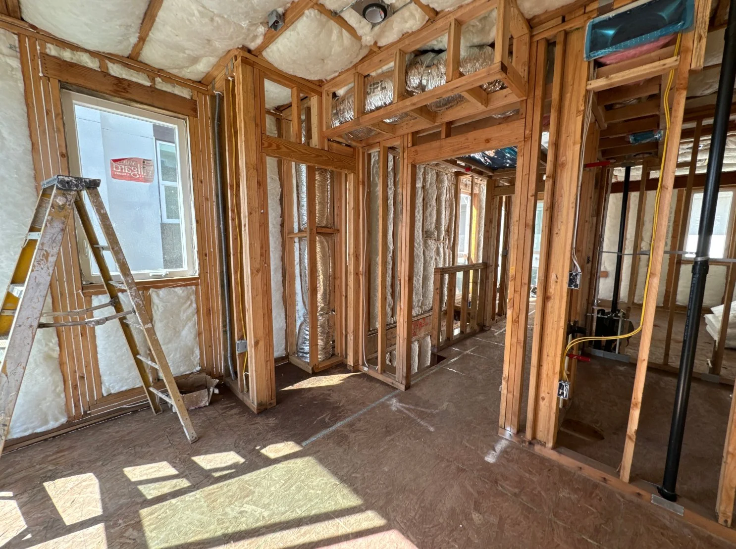 Interior of a house under construction with wooden framing, insulation, an aluminum ladder, and electrical wiring installed.