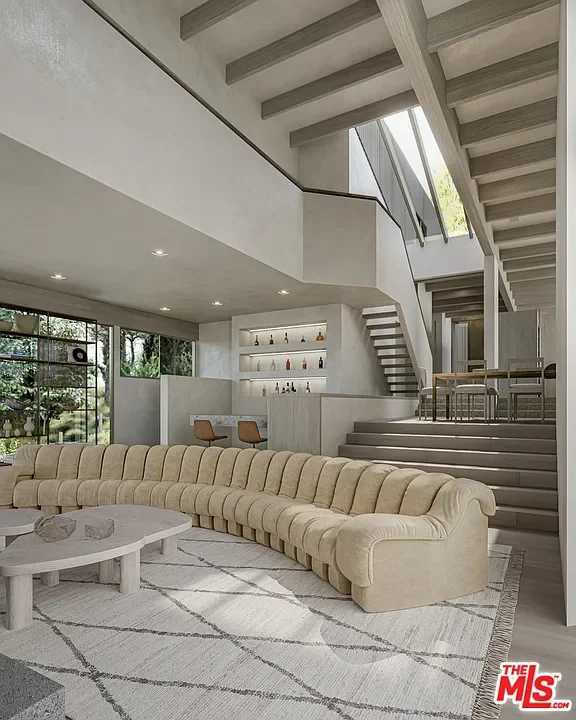 Modern living room with curved beige sofa, glass walls with greenery outside, built-in shelves with bottles, staircase, and open upper level with large windows.