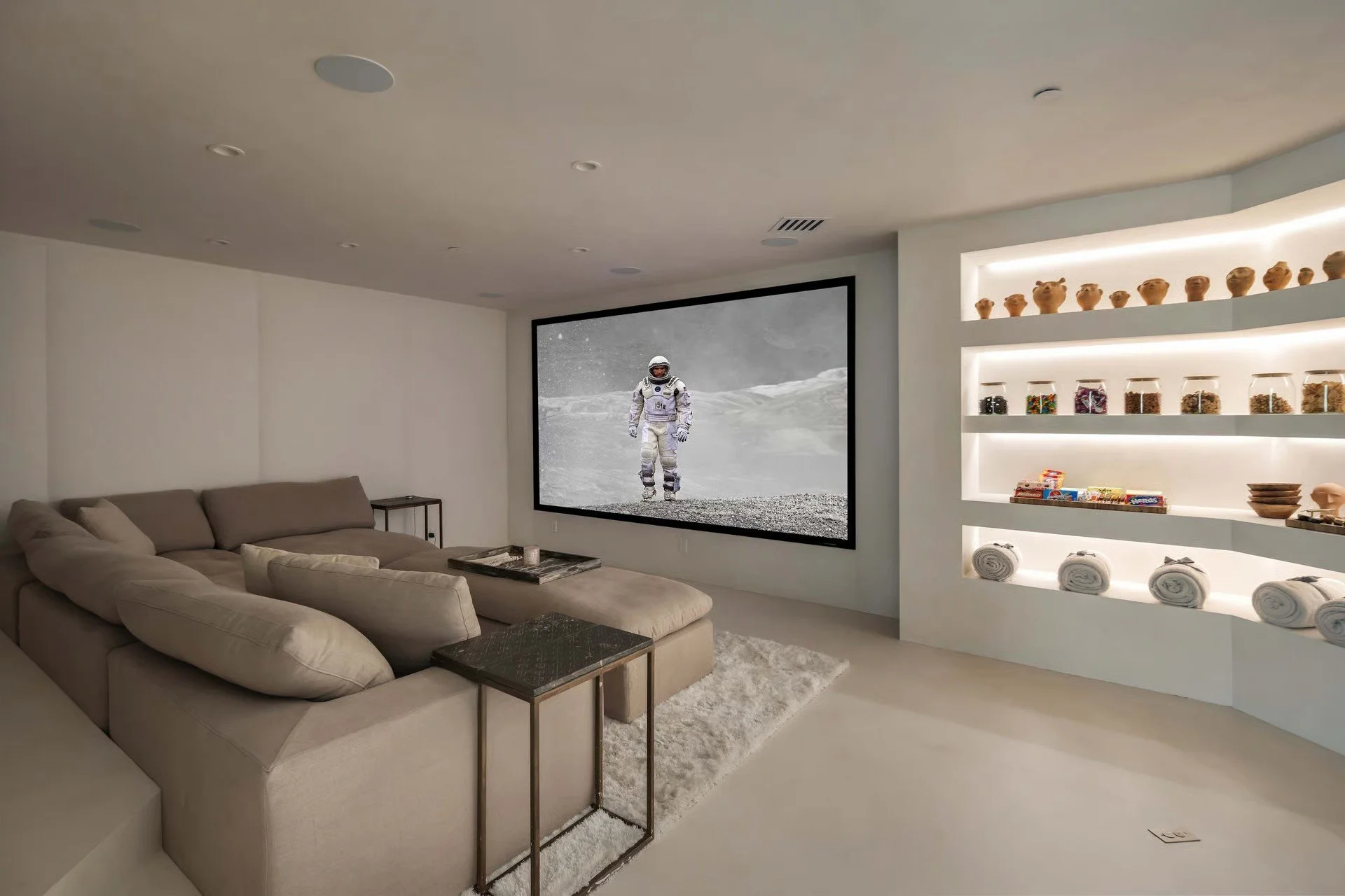 Modern living room with beige sectional sofa, coffee table, wall-mounted TV displaying astronaut in lunar landscape, built-in shelves with pottery, jars, bowls, and rolled towels, and soft lighting.