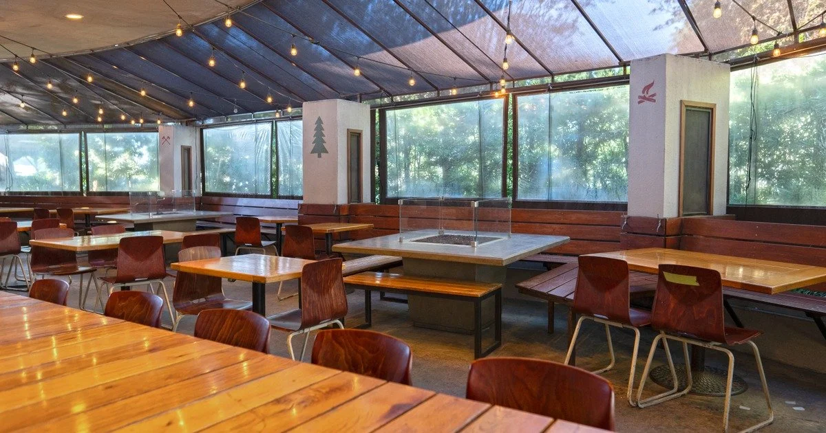 Empty restaurant or cafe with wooden tables and chairs, string lights hanging from the ceiling, large windows with a view of trees outside, and wall decorations.