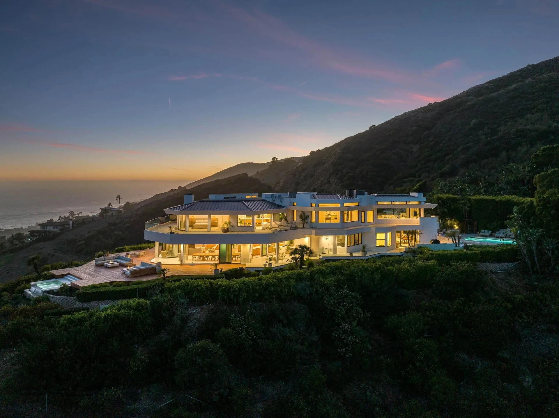 Modern multi-story house on a hillside during sunset with lit interior, outdoor pool, and scenic ocean view in the background.