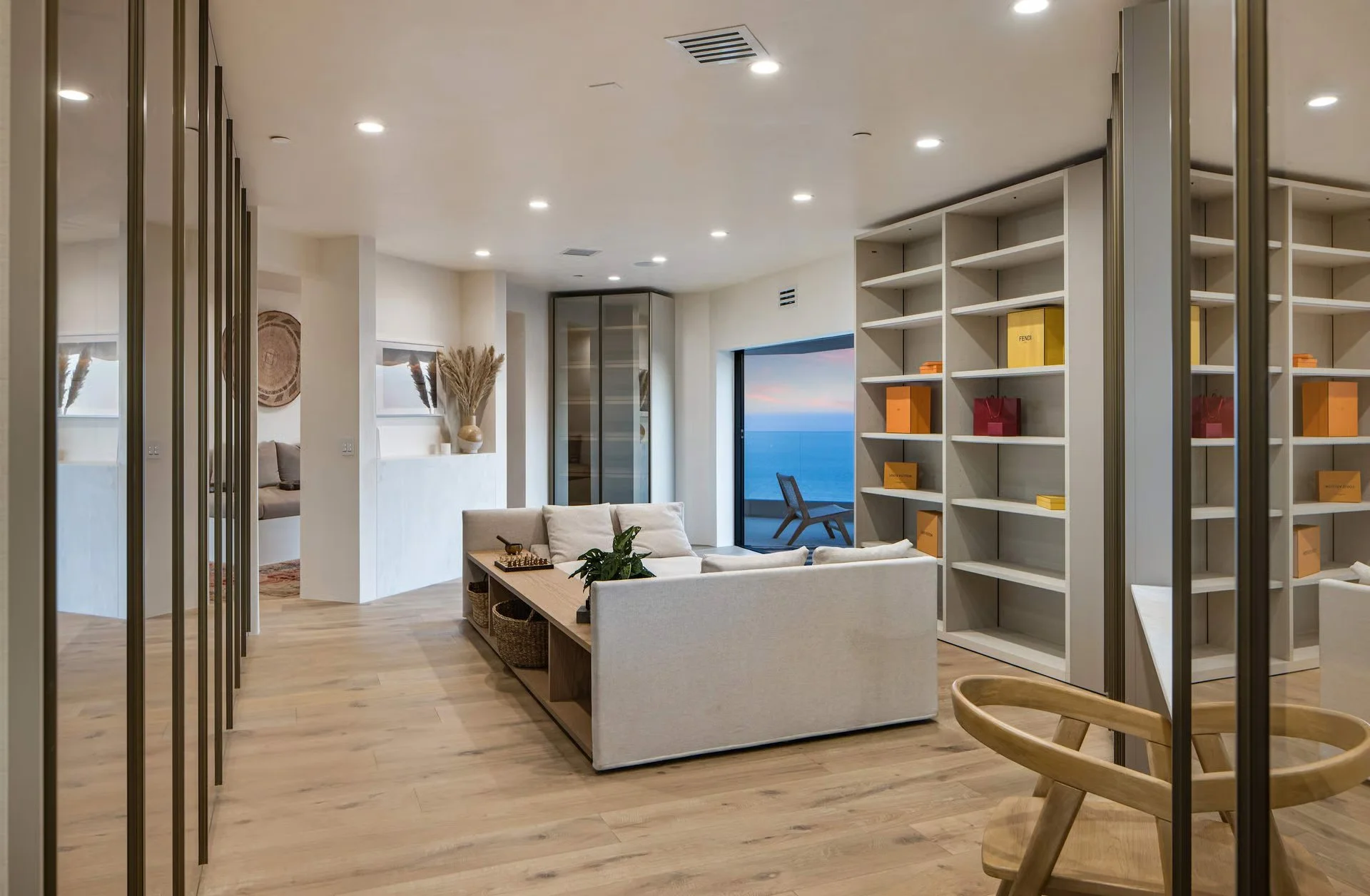 Modern living room with white sofa, open view of the ocean, wooden flooring, built-in shelves with decor and boxes, and a glass door leading to a balcony.