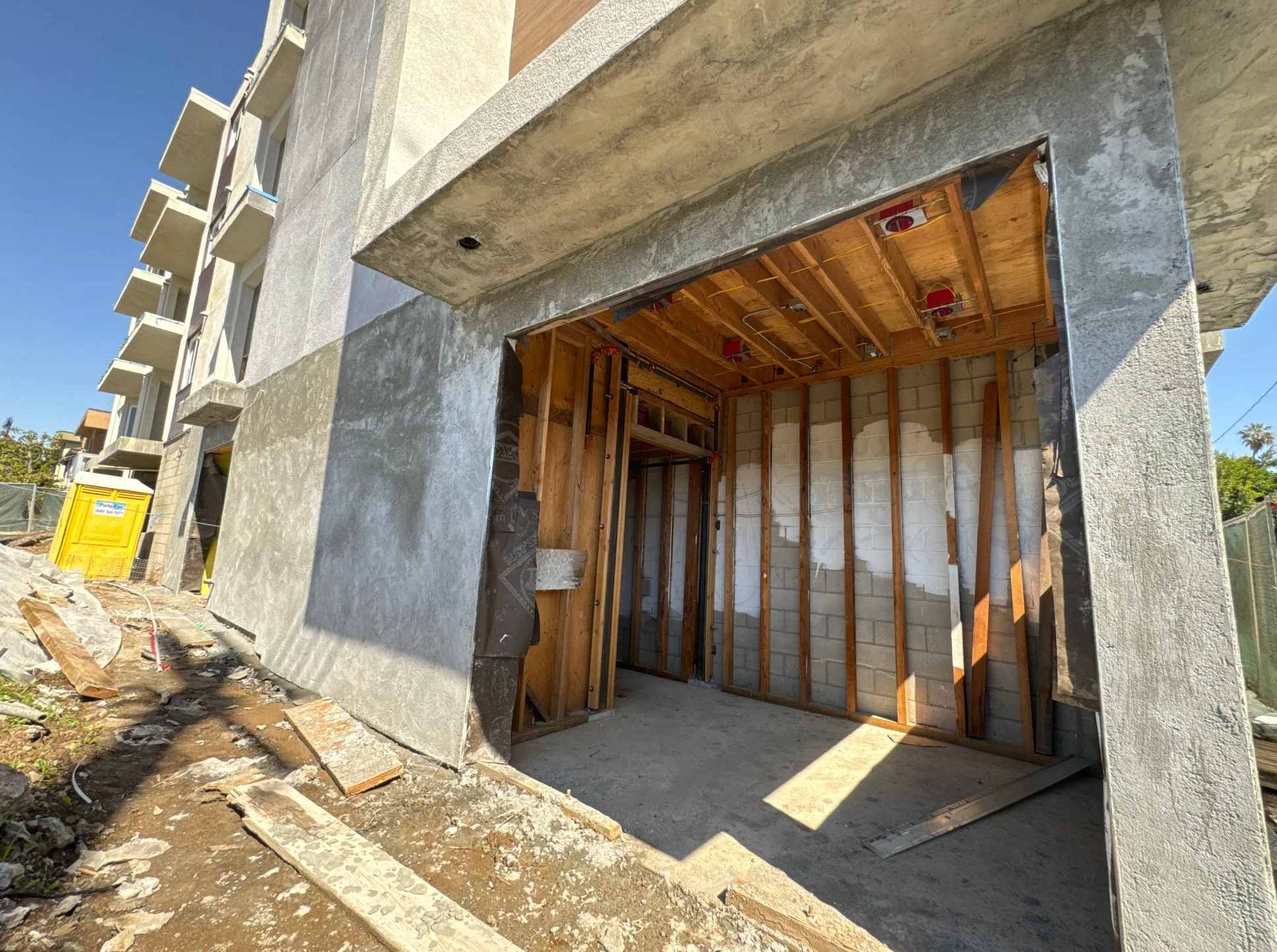 Construction site of a building with an open garage or entryway, showing unfinished interior framing, exposed brick wall, wooden ceiling, and building materials on the ground outside on dirt.