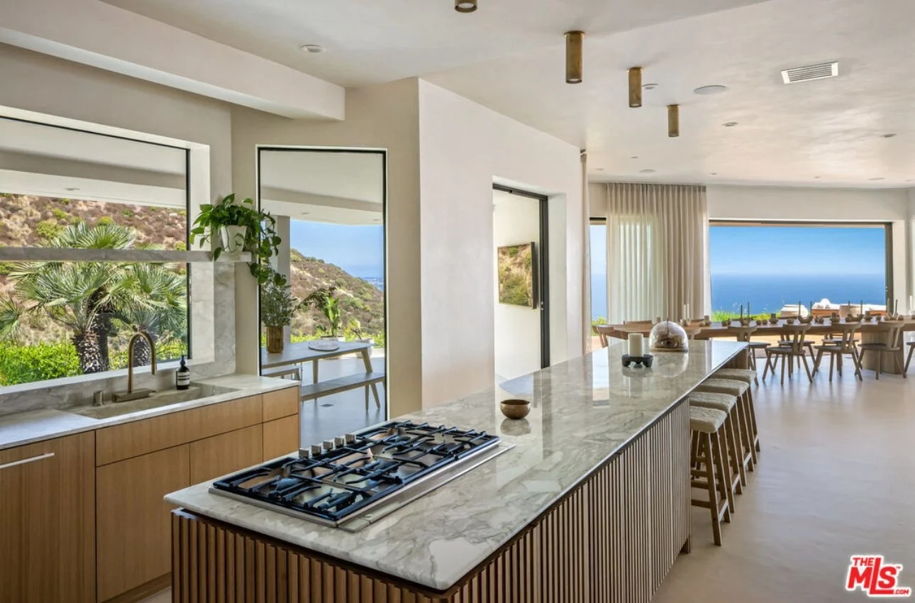 Open-concept kitchen with marble island, gas stove, wooden cabinetry, and large windows showing a hillside and ocean view.