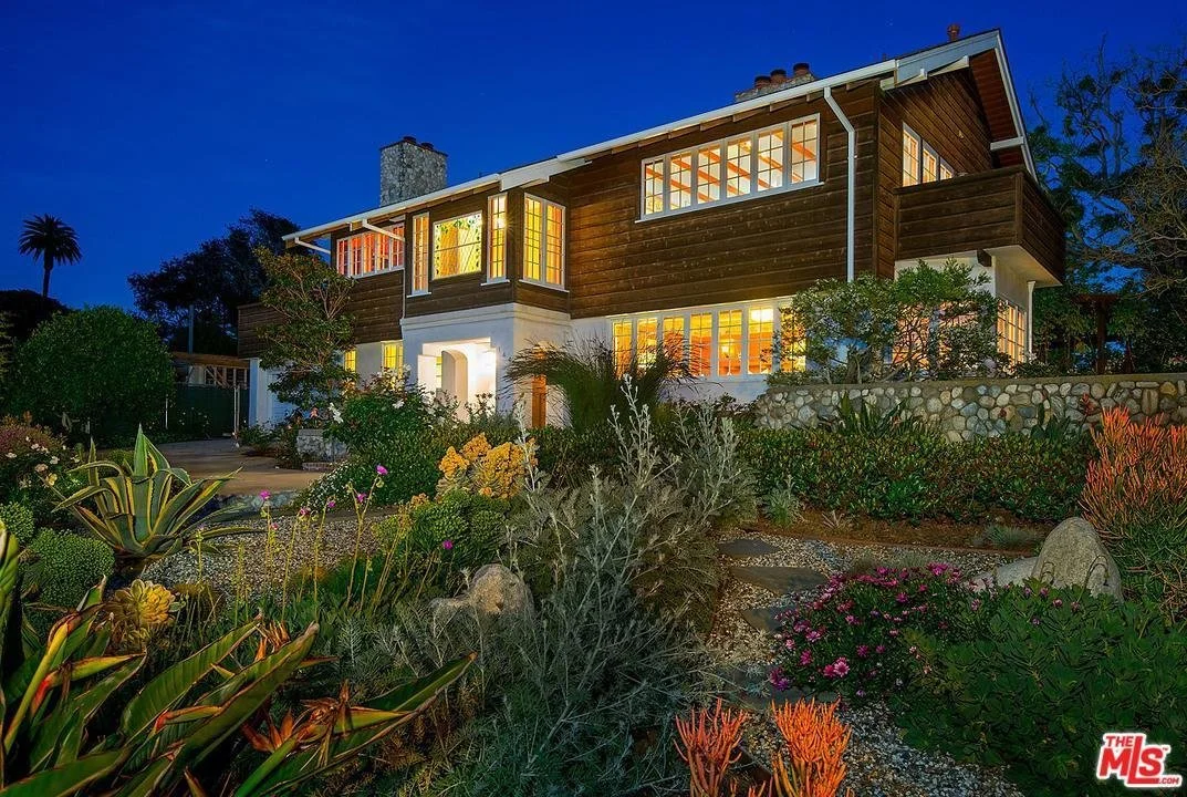 A two-story house with large windows illuminated from within, surrounded by a landscaped garden at dusk. The house features a combination of wood siding and stone foundation, with a prominent chimney and a small balcony on the second floor.