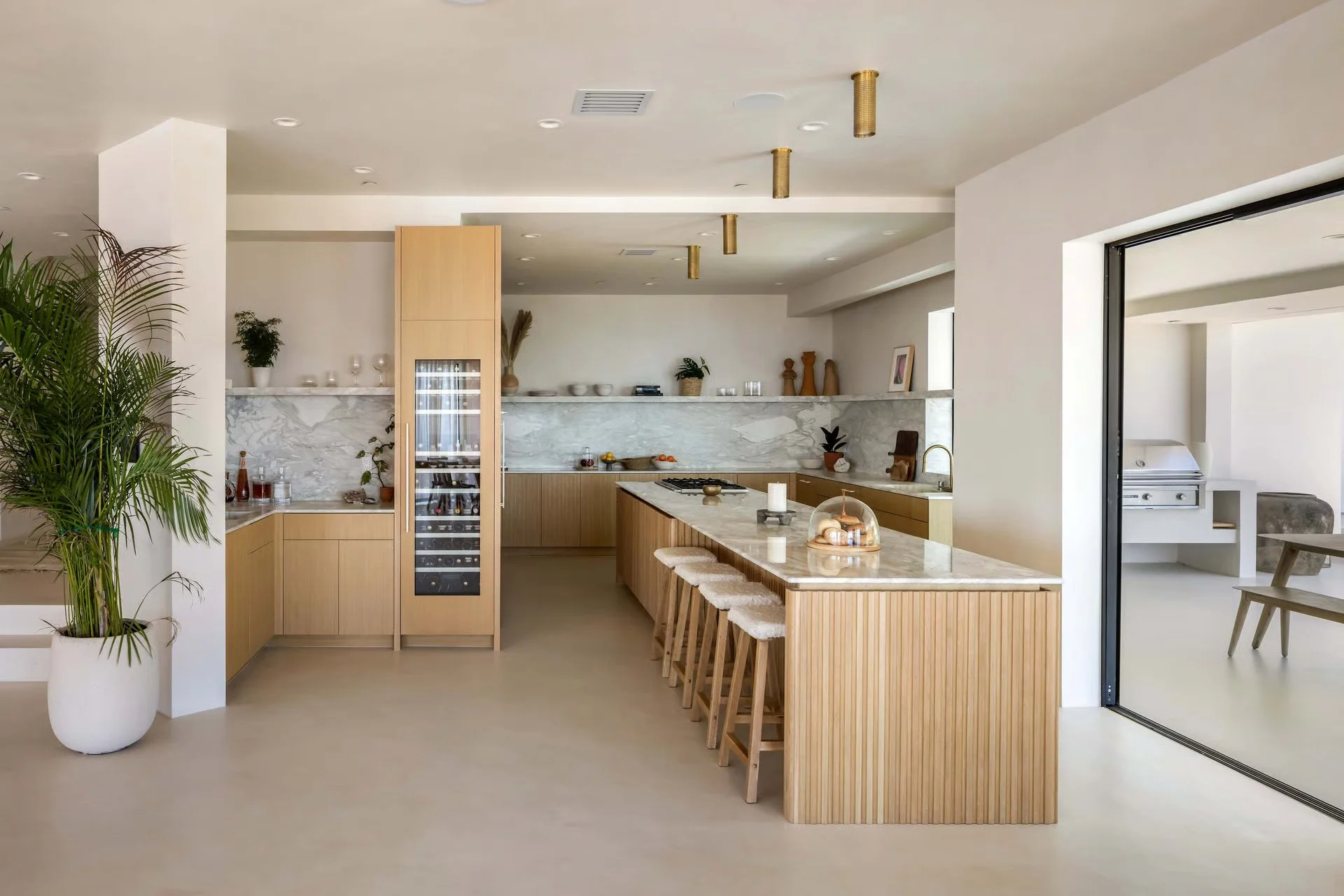 Modern kitchen with wood cabinetry, marble countertops, bar seating, and minimal decor, adjacent to a patio area with seating and a barbecue.