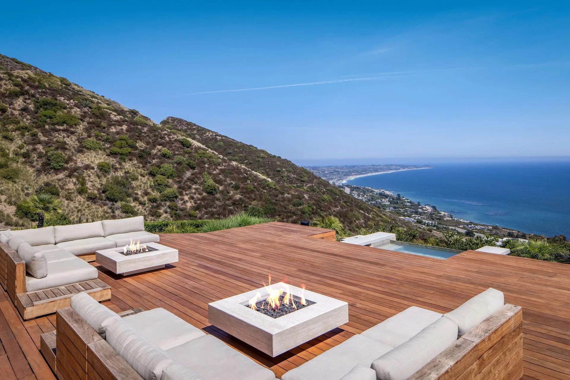 A spacious outdoor wooden deck with white cushioned seating around two modern square fire pits, overlooking a hillside with shrubs and a coastline with an ocean view.