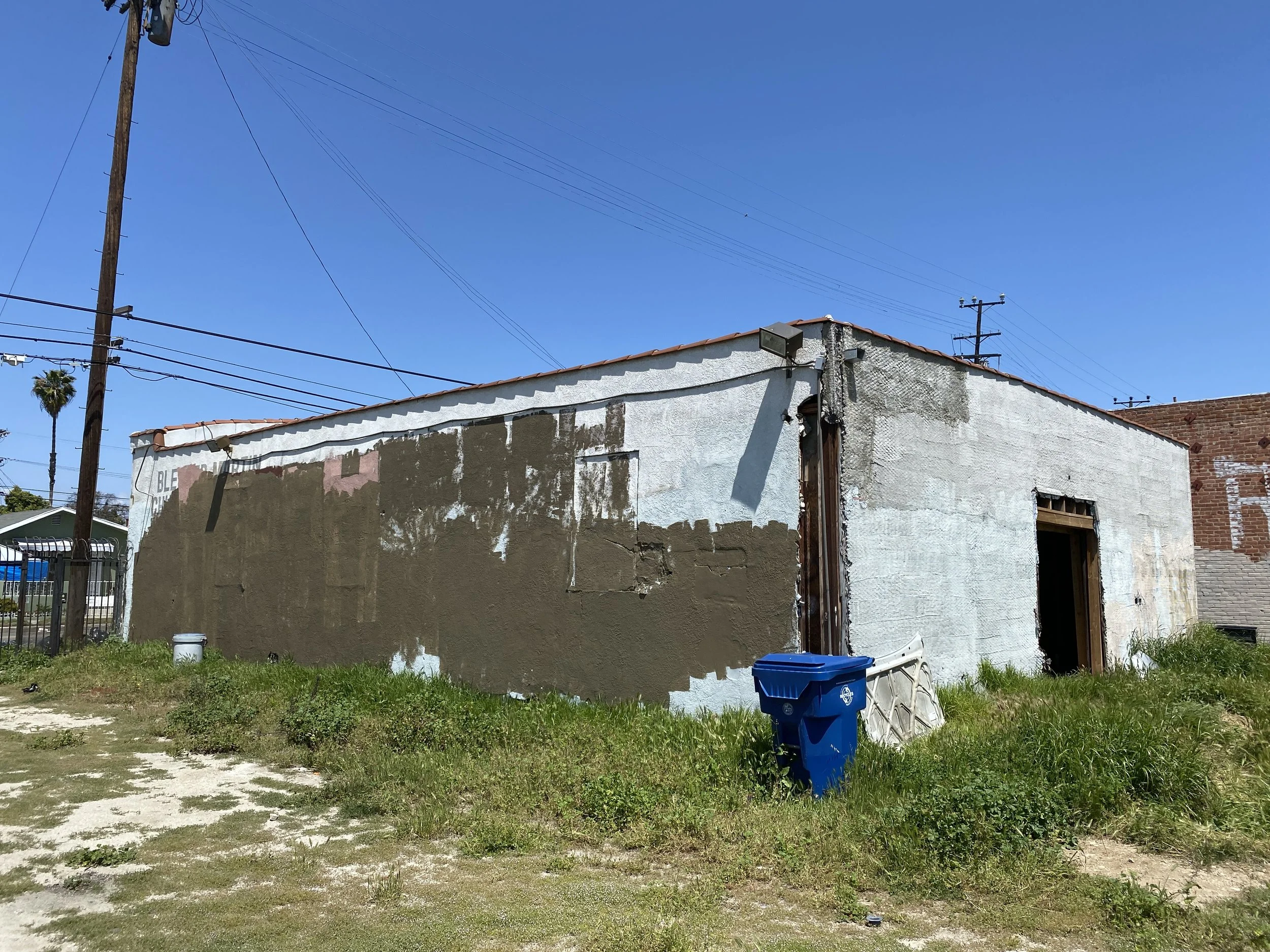 An old building with peeling paint and partially patched wall in front of a clear blue sky. There is a blue trash bin and some grass and dirt in the foreground.