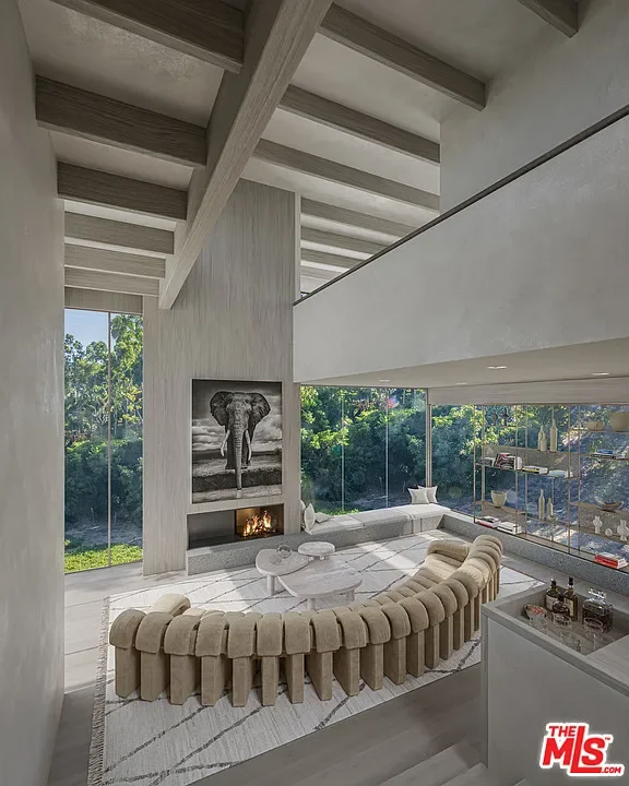 Modern living room with a curved beige velvet sofa, white coffee table, and floor-to-ceiling windows showing lush greenery outside. There's a fireplace with an elephant photo above it and open shelving on the right.