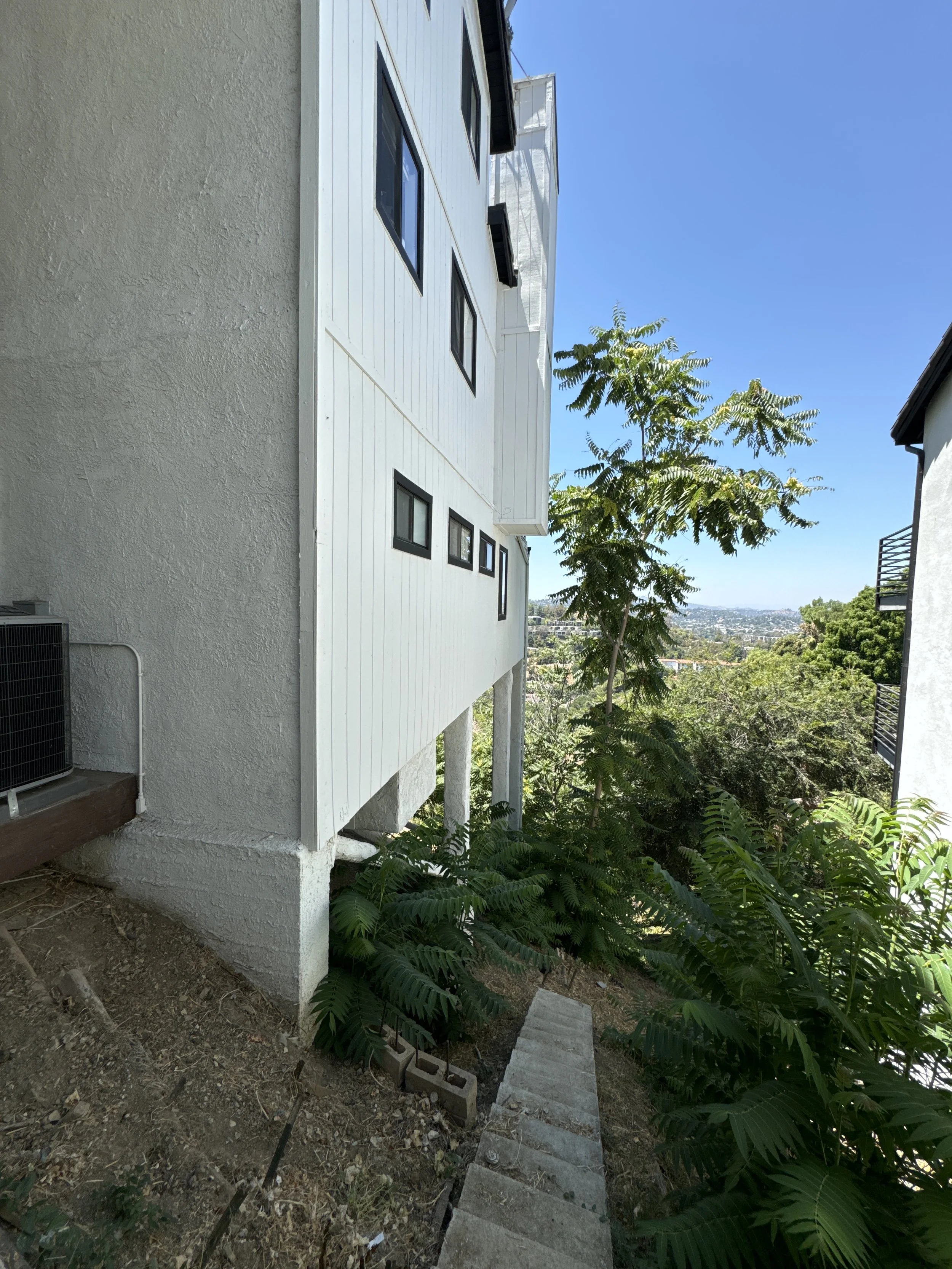 View of the side of a modern white building with black window frames, a concrete pathway leading down a hill, dense green foliage, and a clear blue sky in the background.