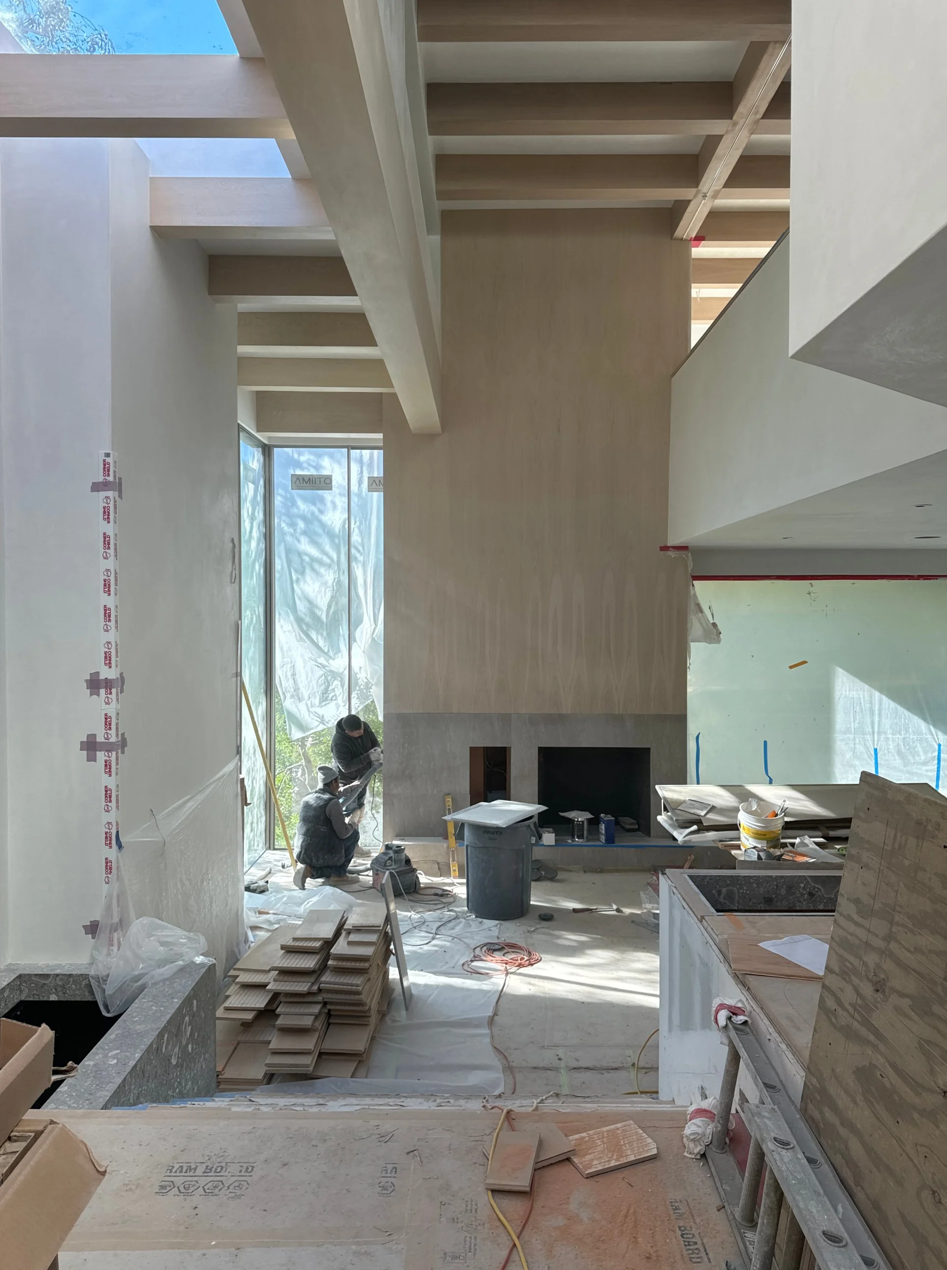 Construction workers installing fixtures in a modern, spacious home interior with large windows, a high ceiling, and ongoing tiling and finishing work.