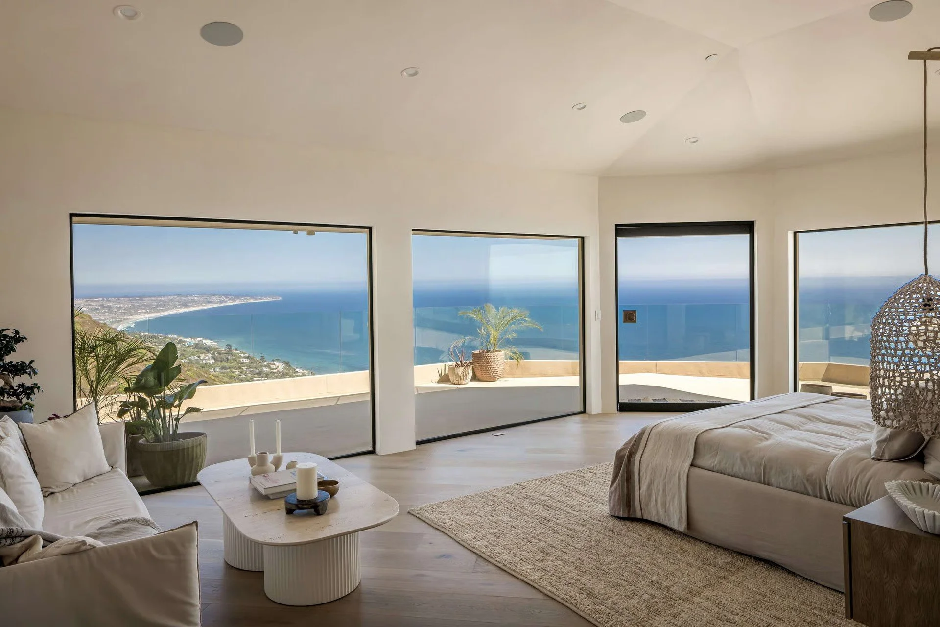 Bright bedroom with large windows showcasing a coastal view, a white bed, a woven pendant light, and neutral decor.