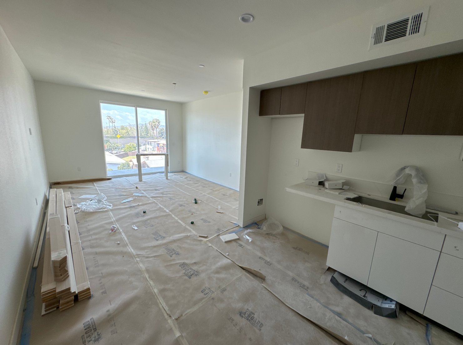 An unfinished living room with large windows, white walls, and a patio door leading outside. The floor is covered with protective paper and construction debris. A kitchen area with white cabinetry and dark wood upper cabinets is visible on the right.
