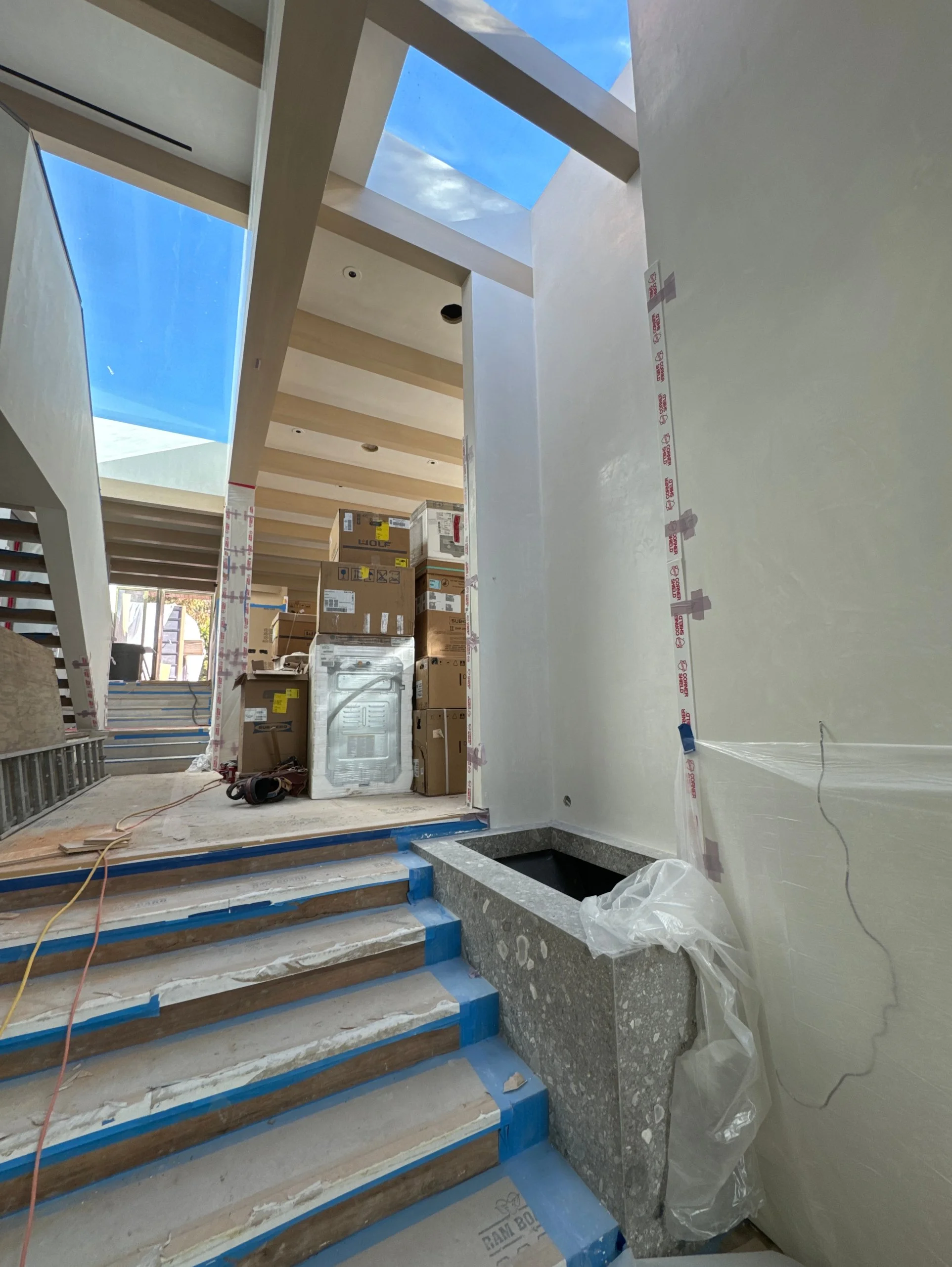 Interior of a modern house under construction, featuring a staircase with protective blue tape, boxes of building materials, and partially finished walls under a skylight ceiling.