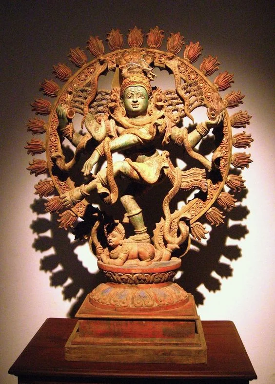 Wooden sculpture of a multi-armed deity with a crown, surrounded by a circular halo of flames, standing on a platform with a lion figure at the base, displayed on a wooden table.