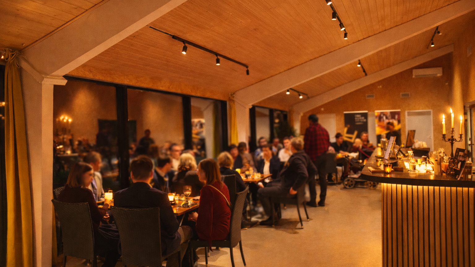 Indoor restaurant or event space filled with people seated at tables, lit by candlelight, with a bar area on the right and windows behind the diners; the ceiling is wooden with track lighting.