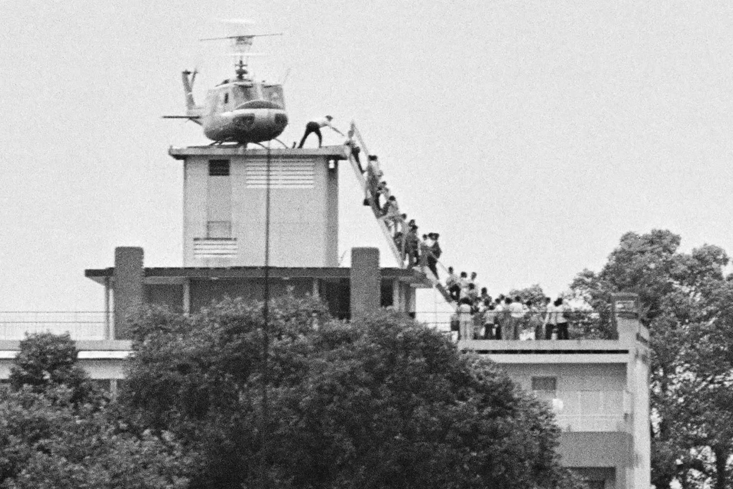 A C.I.A. employee evacuates South Vietnamese refugees into a helicopter from an apartment rooftop during the Fall of Saigon (1975). Photo by Hugh Van Es and via History (color-corrected and cropped).