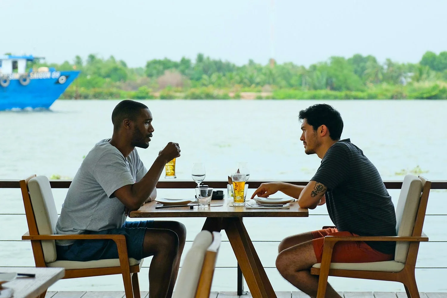 Right to left: Kit (Henry Golding) and Lewis (Parker Sawyers) dine at The Deck Saigon, image still from Monsoon, directed by Hong Khaou (2019). Via MoMA (color-corrected and cropped).