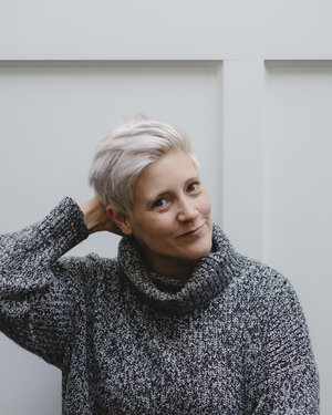 a color picture of emily m danforth, taken from her website and credited to chris mongeau. she is a white woman with short white hair and dark eyes who is smiling at the viewer. she is wearing a grey turtleneck, and is rubbing the back of her head, as if bashful.