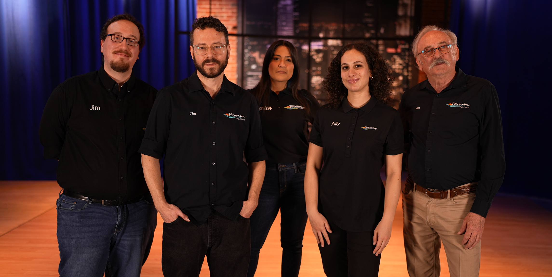 Group of five people standing together on a stage with a wooden floor and dark blue curtains, wearing matching black shirts with logos.