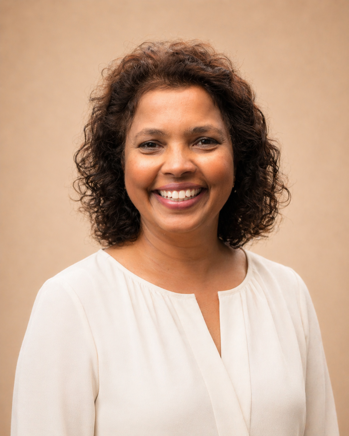 Smiling woman with curly hair wearing a black dress with white floral pattern, standing outdoors in front of green foliage.
